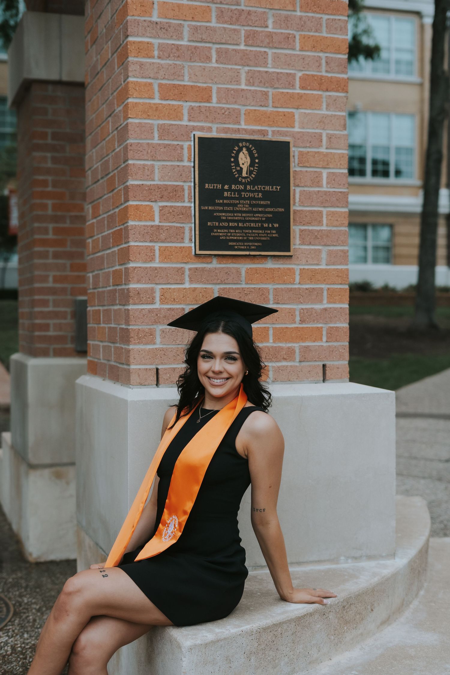 Graduate in cap and gown with orange stole sits on brick ledge by campus plaque.