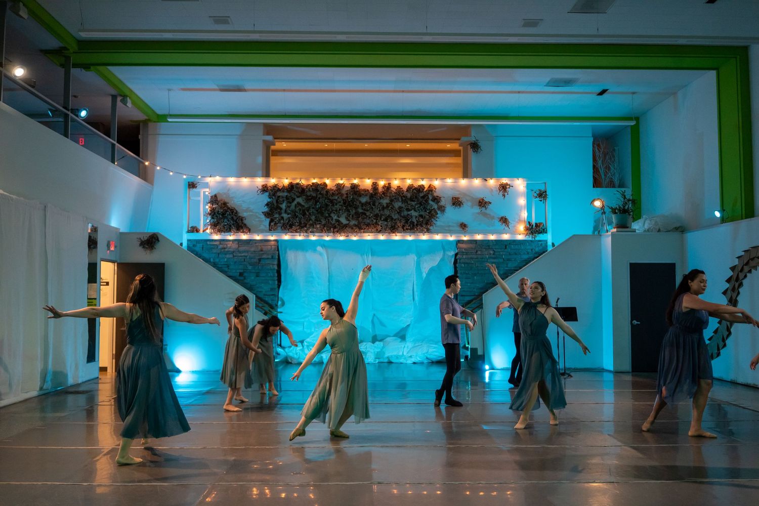 Dancers in flowing gray dresses perform gracefully in a teal-lit hall with a floral backdrop and string lights.