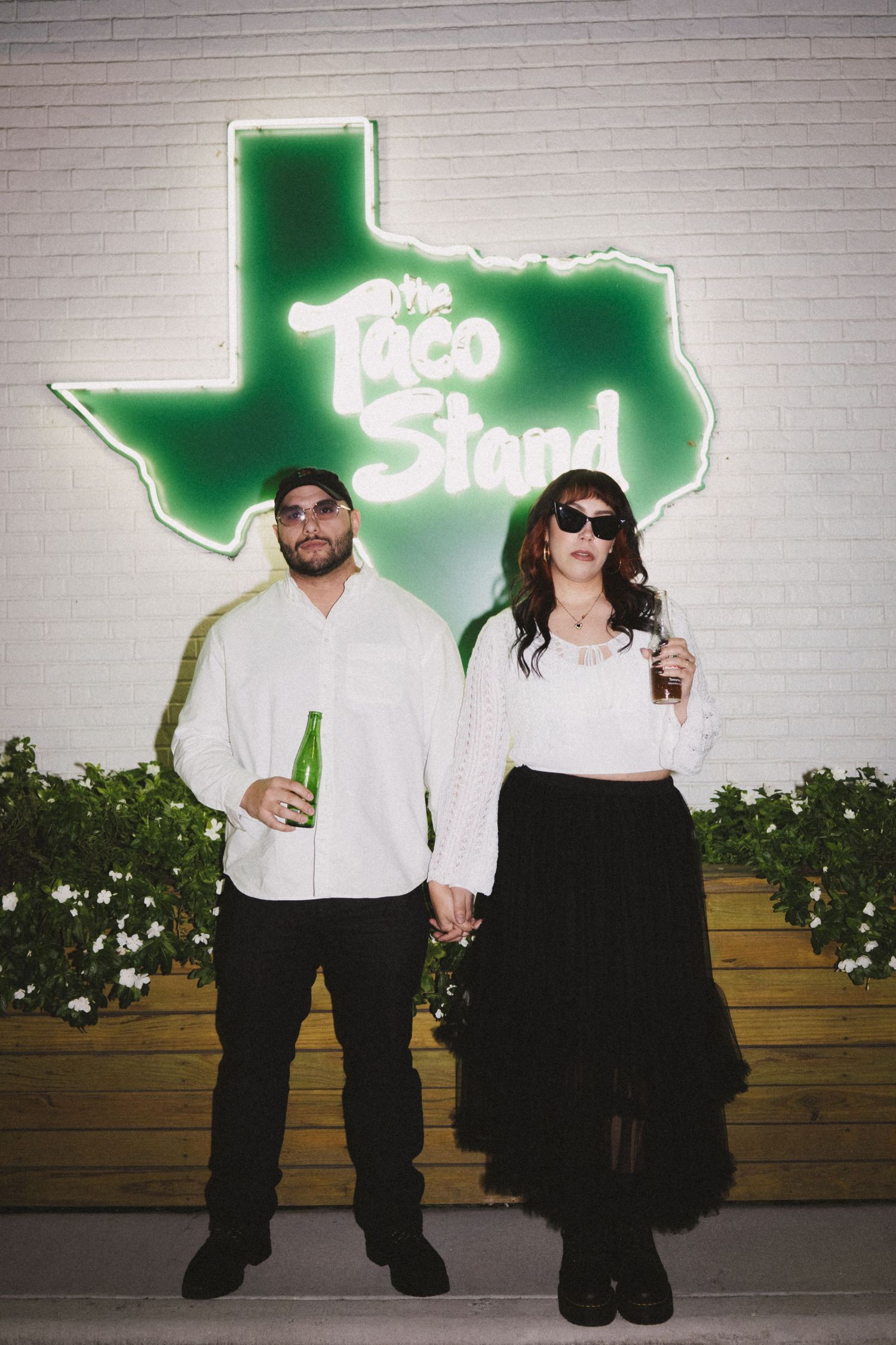 Couple posing in front of a green neon Texas Taco Stand sign, dressed in black and white, holding green bottles.
