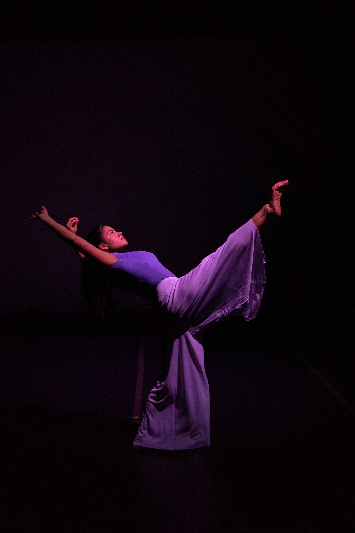 A dancer in purple performs a dramatic pose with leg raised high under stage lighting against a dark background.