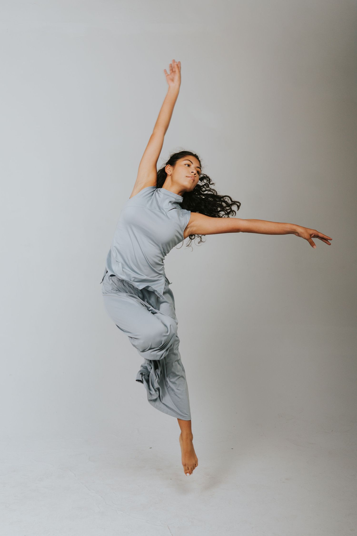 A dancer leaps gracefully in a gray outfit, arms extended, against a light background.