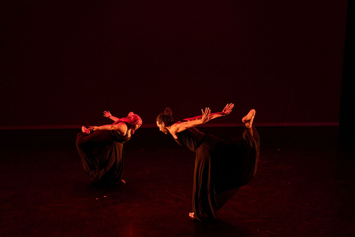 Dancers perform in dramatic red stage lighting, their flowing dark costumes creating elegant silhouettes on a dimly lit stage.