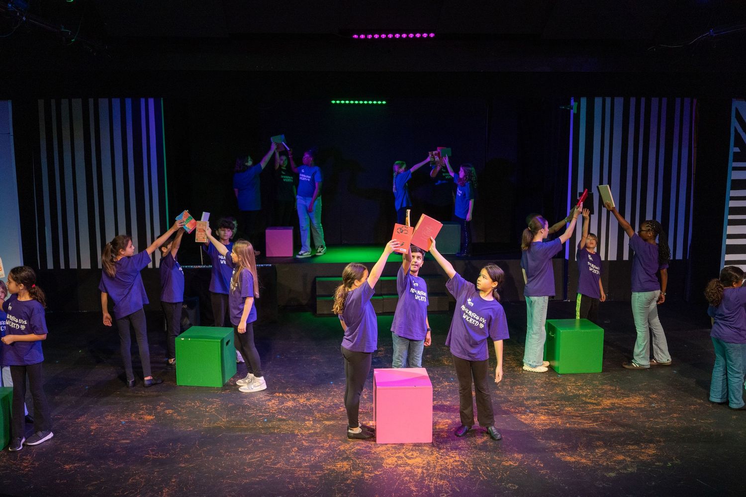 Children in purple shirts perform on stage with colorful props and neon lighting during a school theater production.