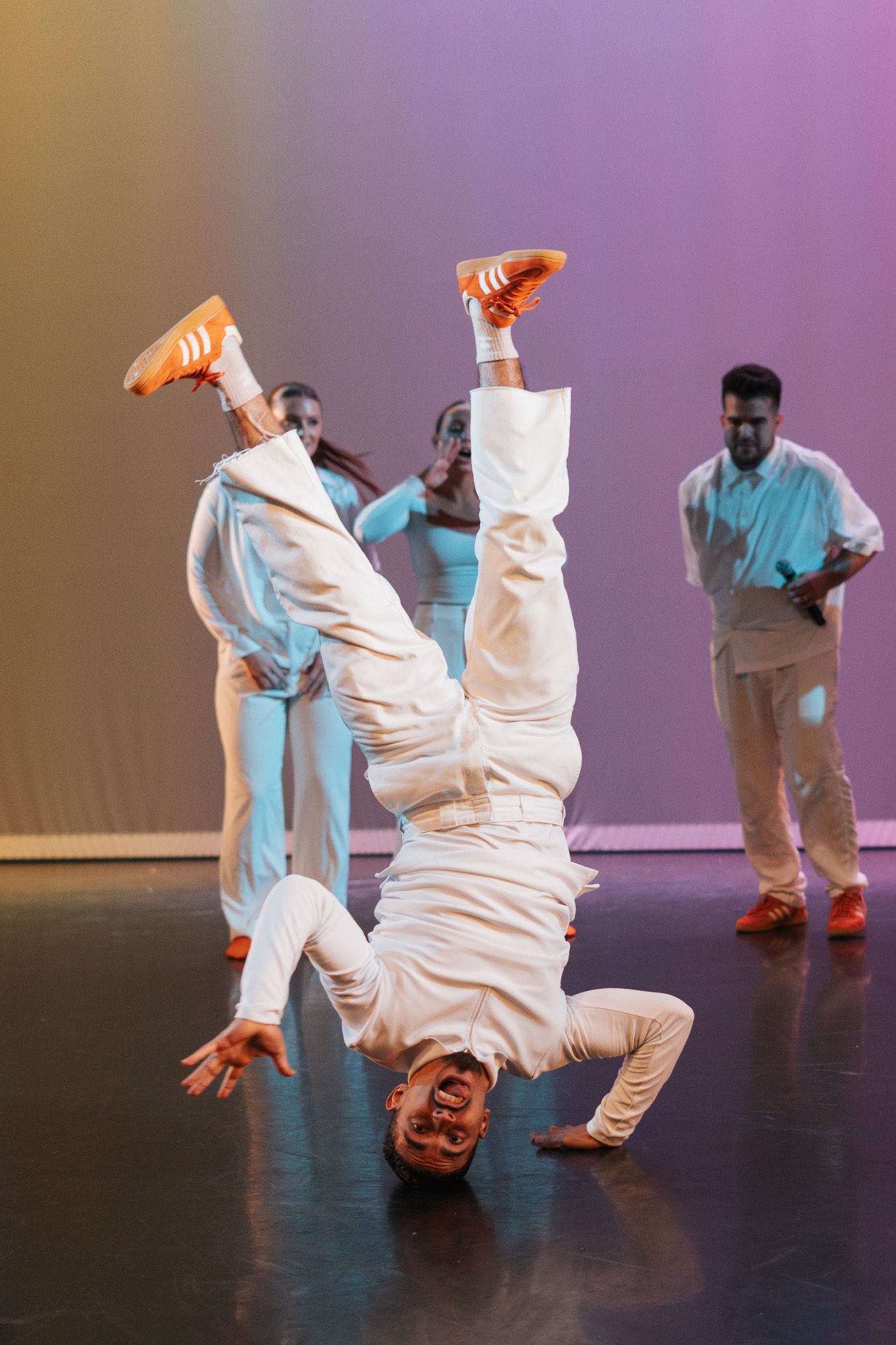 Dancer performs impressive headstand breakdancing move in white outfit while others watch during stage performance.