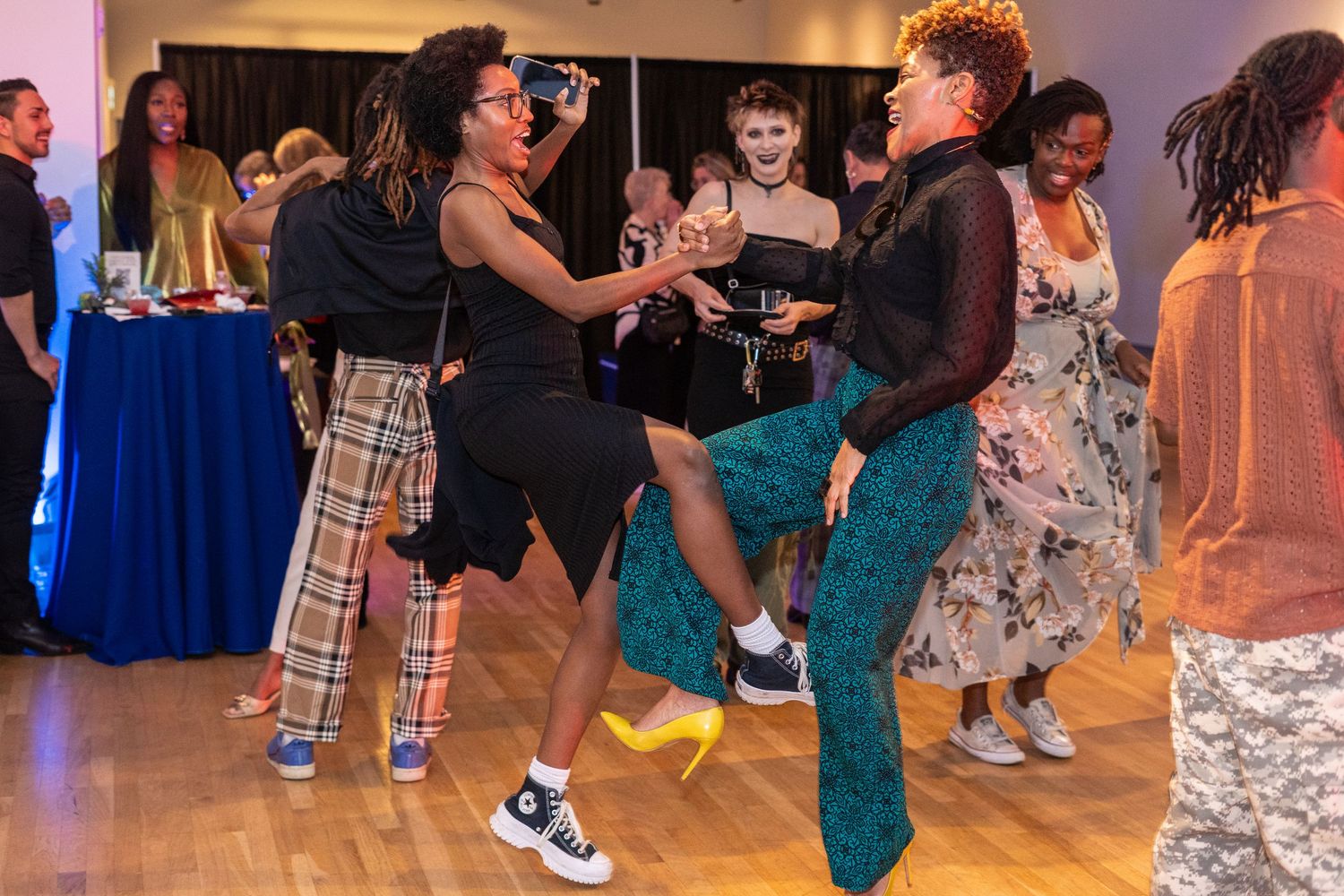 People dancing energetically at an indoor event on a wooden dance floor, dressed in colorful outfits.