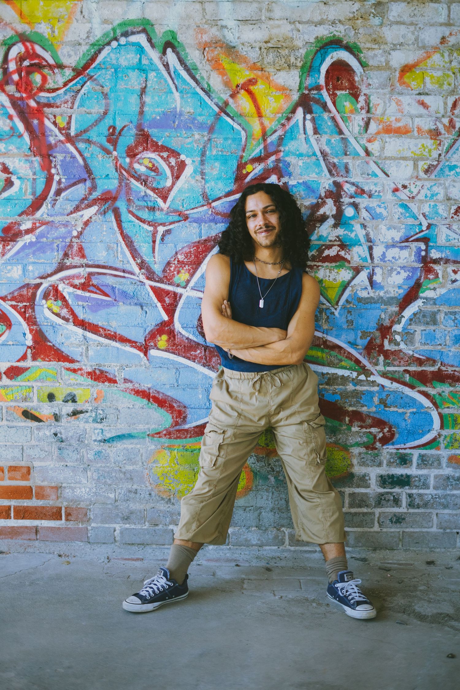 Person posing confidently against vibrant graffiti wall wearing tank top and cargo pants.