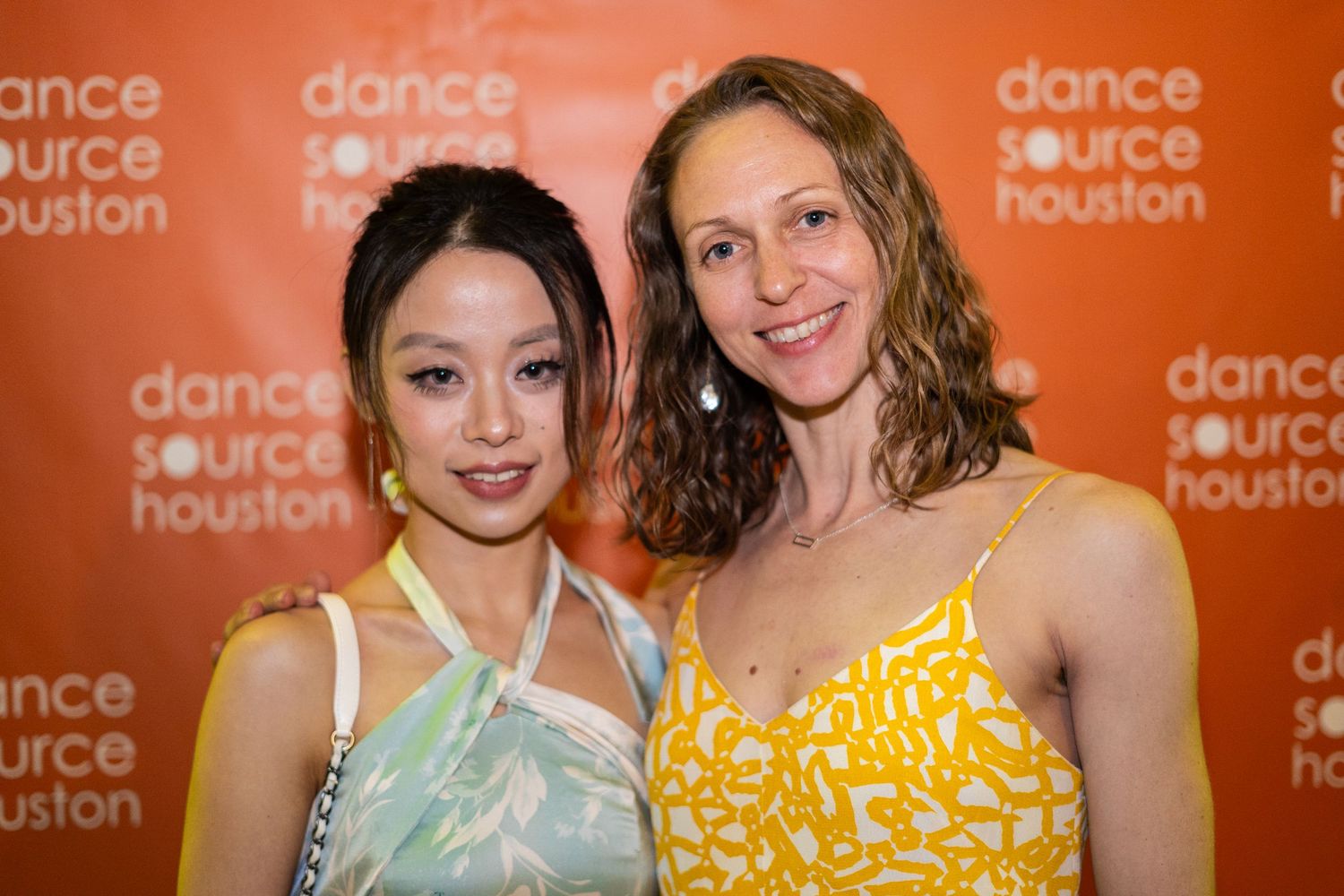 Two dancers pose in colorful dresses against an orange backdrop at a dance event.