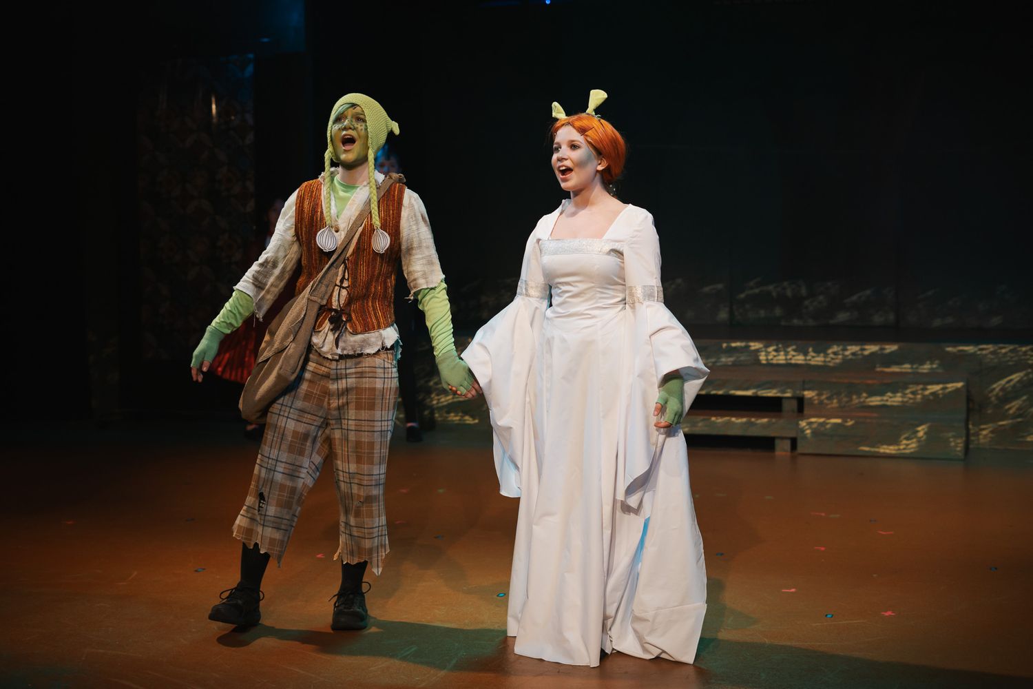 Two actors in Shrek costumes perform on stage, one dressed as Shrek and one as Princess Fiona in a white gown.