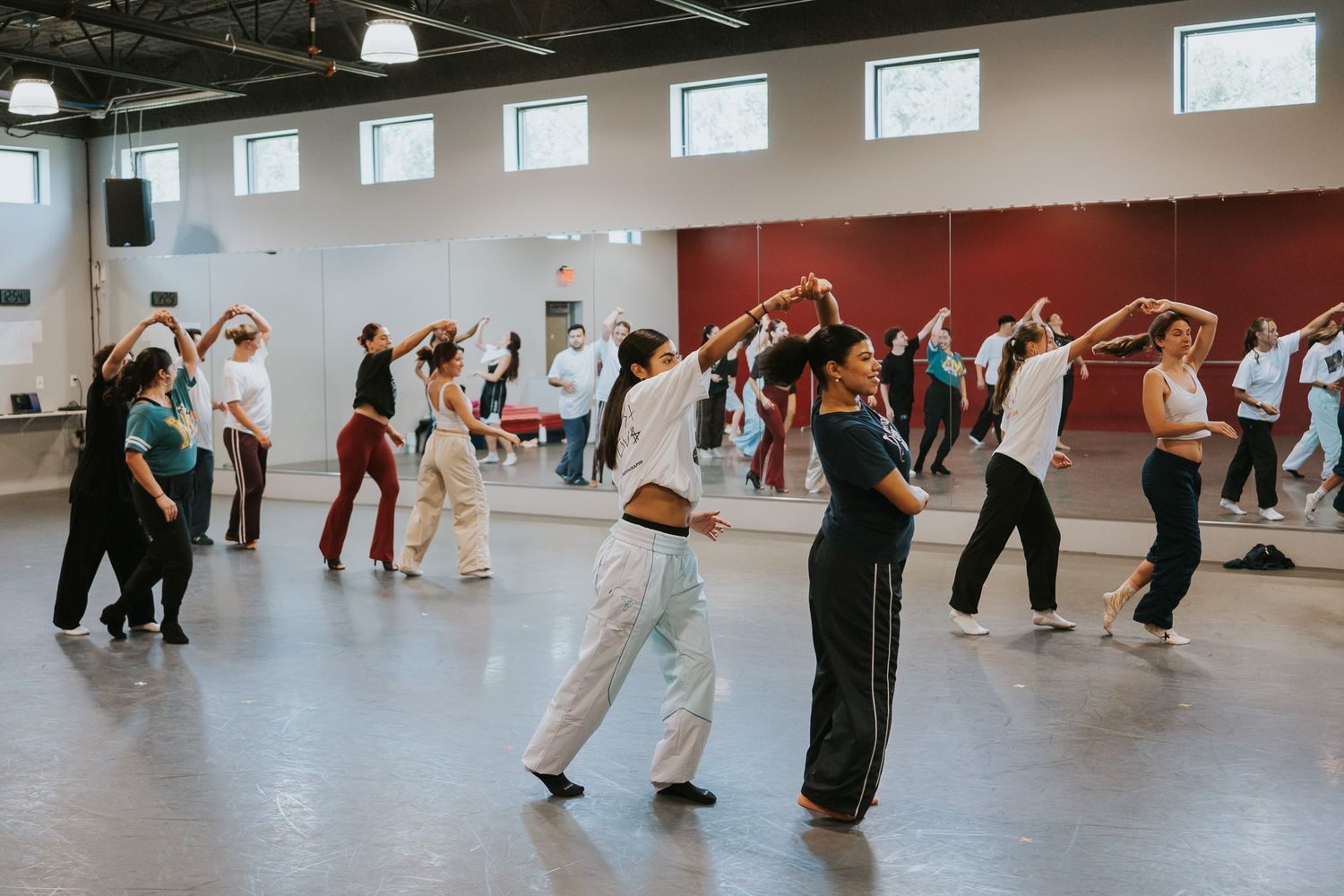 A large group of dancers practice choreography in a spacious studio with high ceilings.