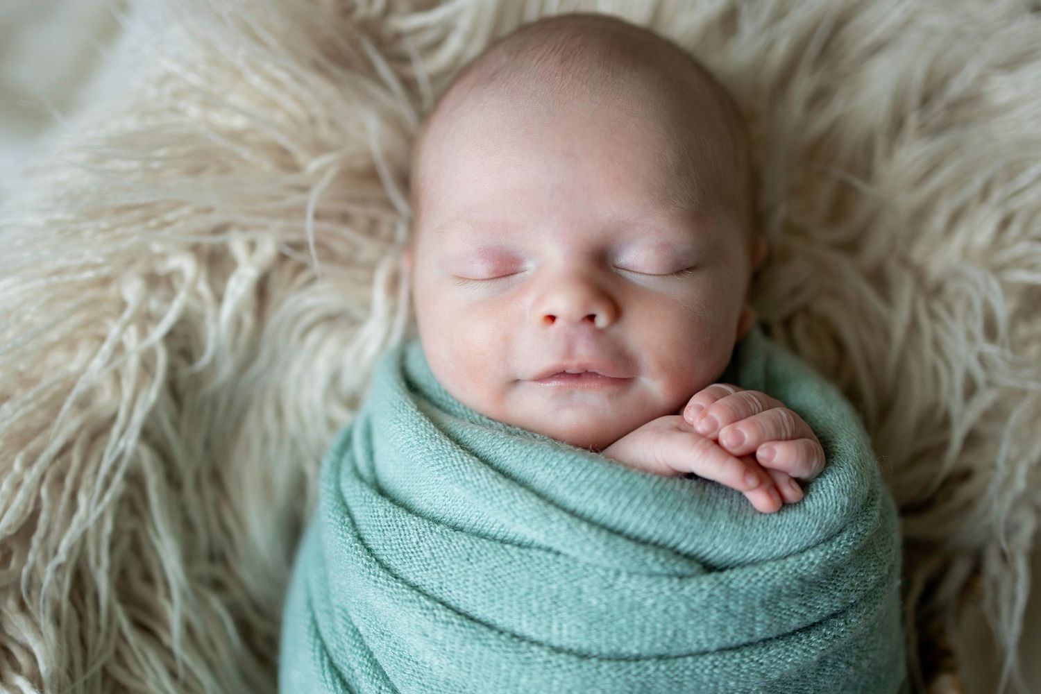 Newborn baby swaddled in a soft green wrap, sleeping peacefully on a fluffy cream blanket.