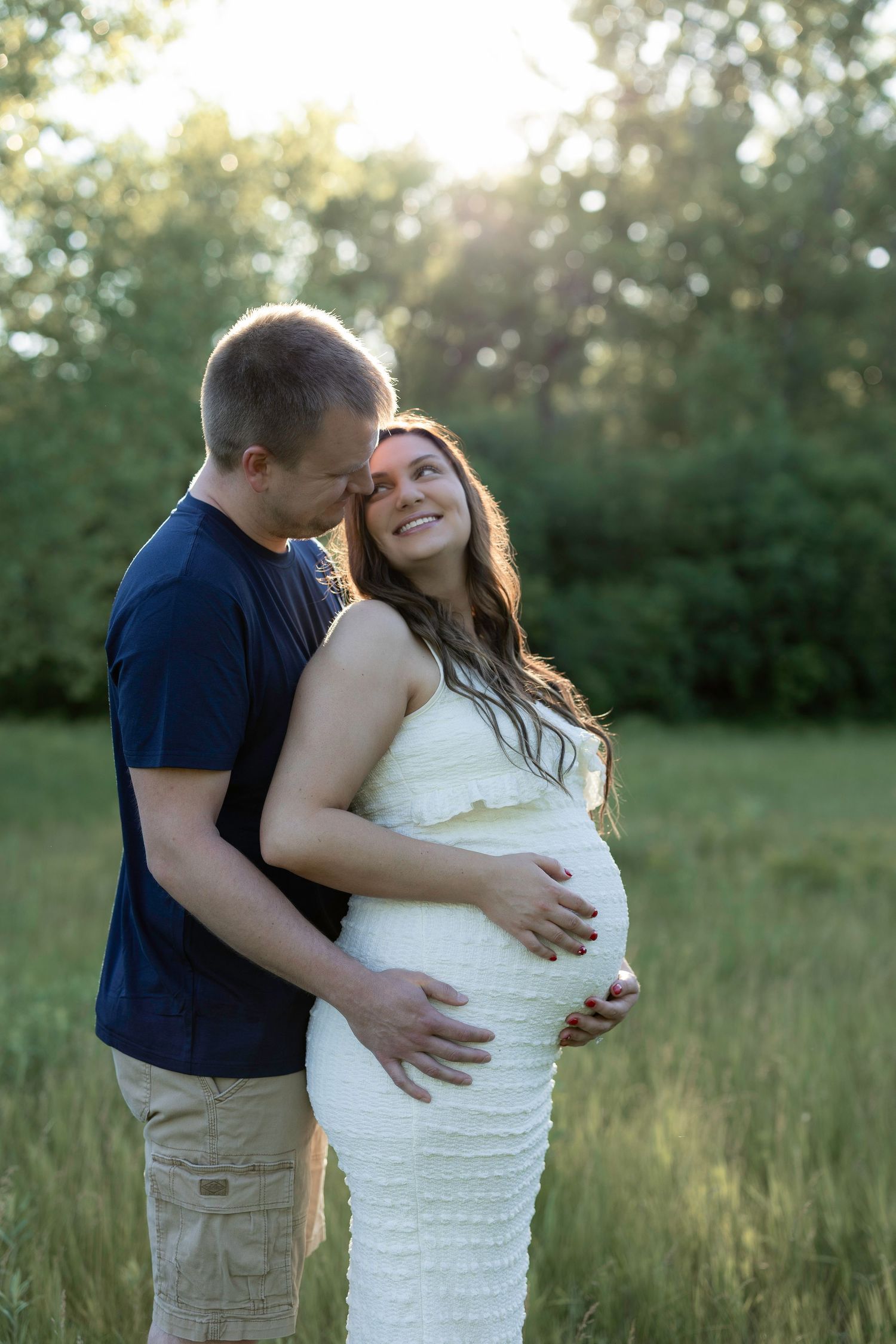 Couple sharing a tender moment outdoors, pregnant woman in white dress smiling up at partner in sunlit green field.