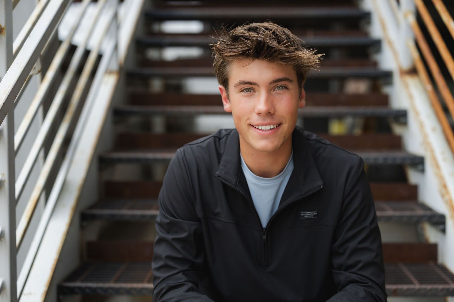 Young person wearing dark quarter-zip sweater sits smiling on wooden stairs with metal railings.