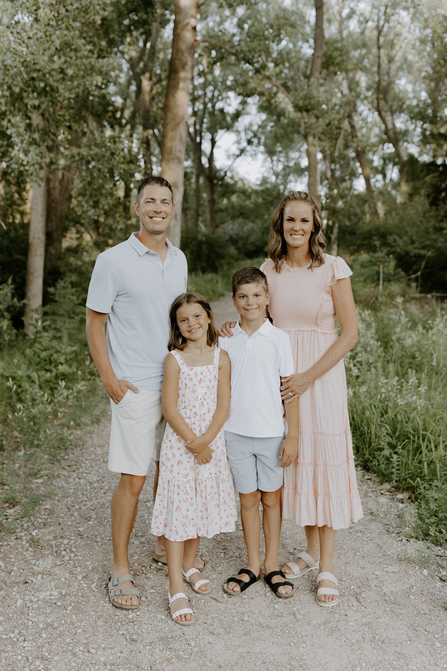 A family wearing pastel colors stands together on a gravel path surrounded by green trees and natural foliage.