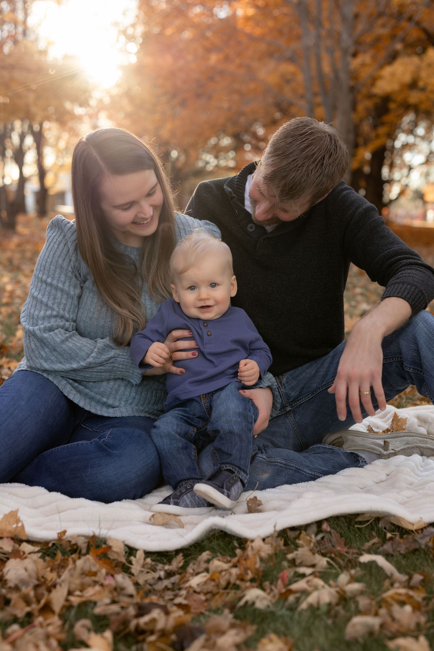 Family of three sitting on blanket in autumn park, parents smiling at baby in purple outfit with golden fall foliage.