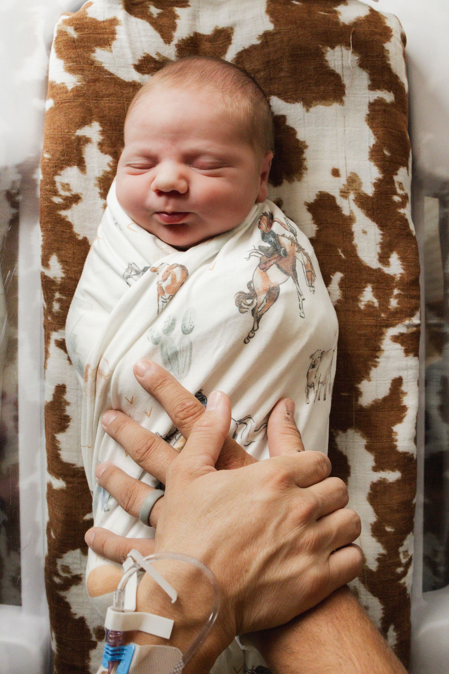 Newborn baby swaddled in cowboy print blanket smiling in hospital bassinet with parent's hand resting gently on chest.
