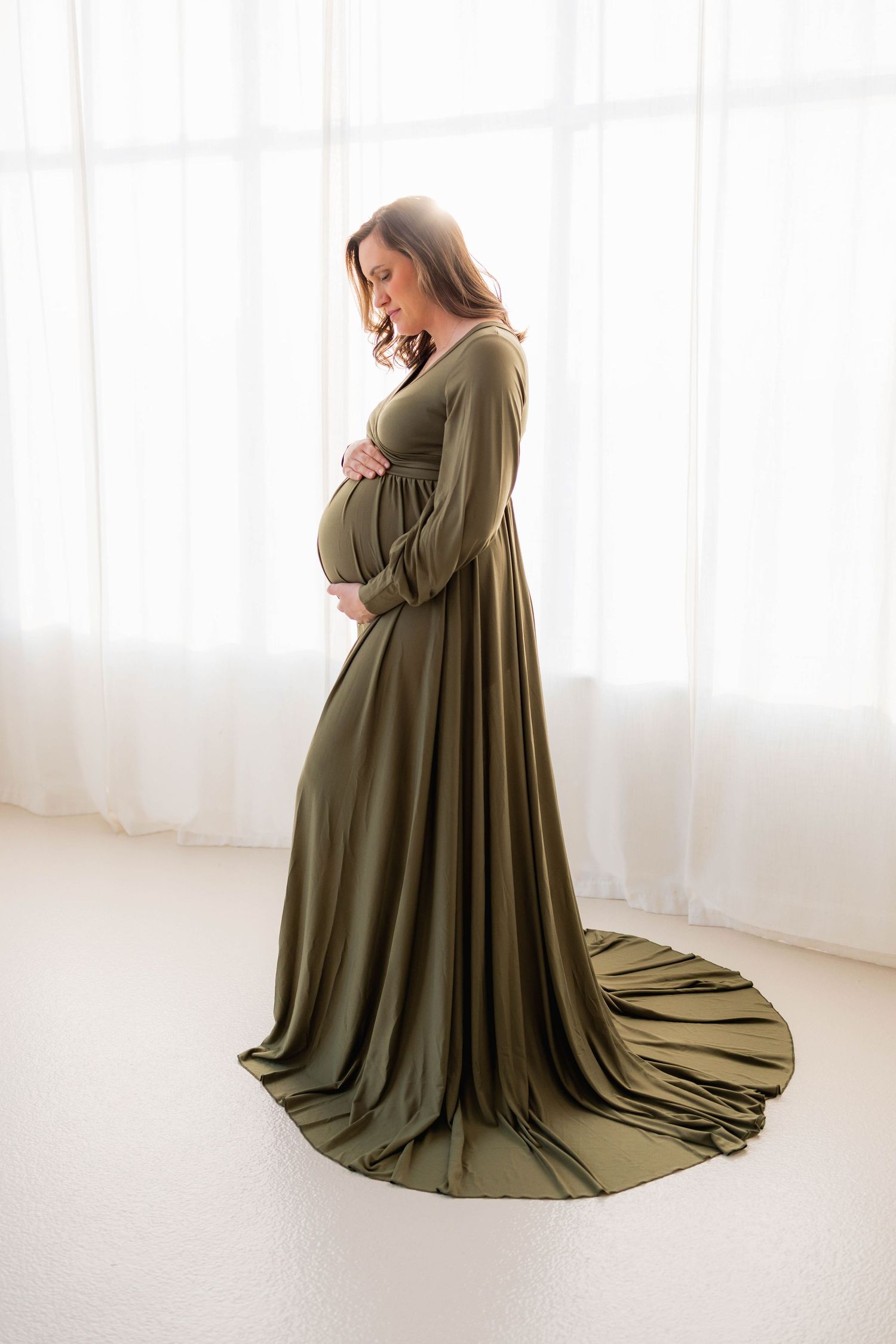 Expectant mother in olive green flowing gown poses by bright window.