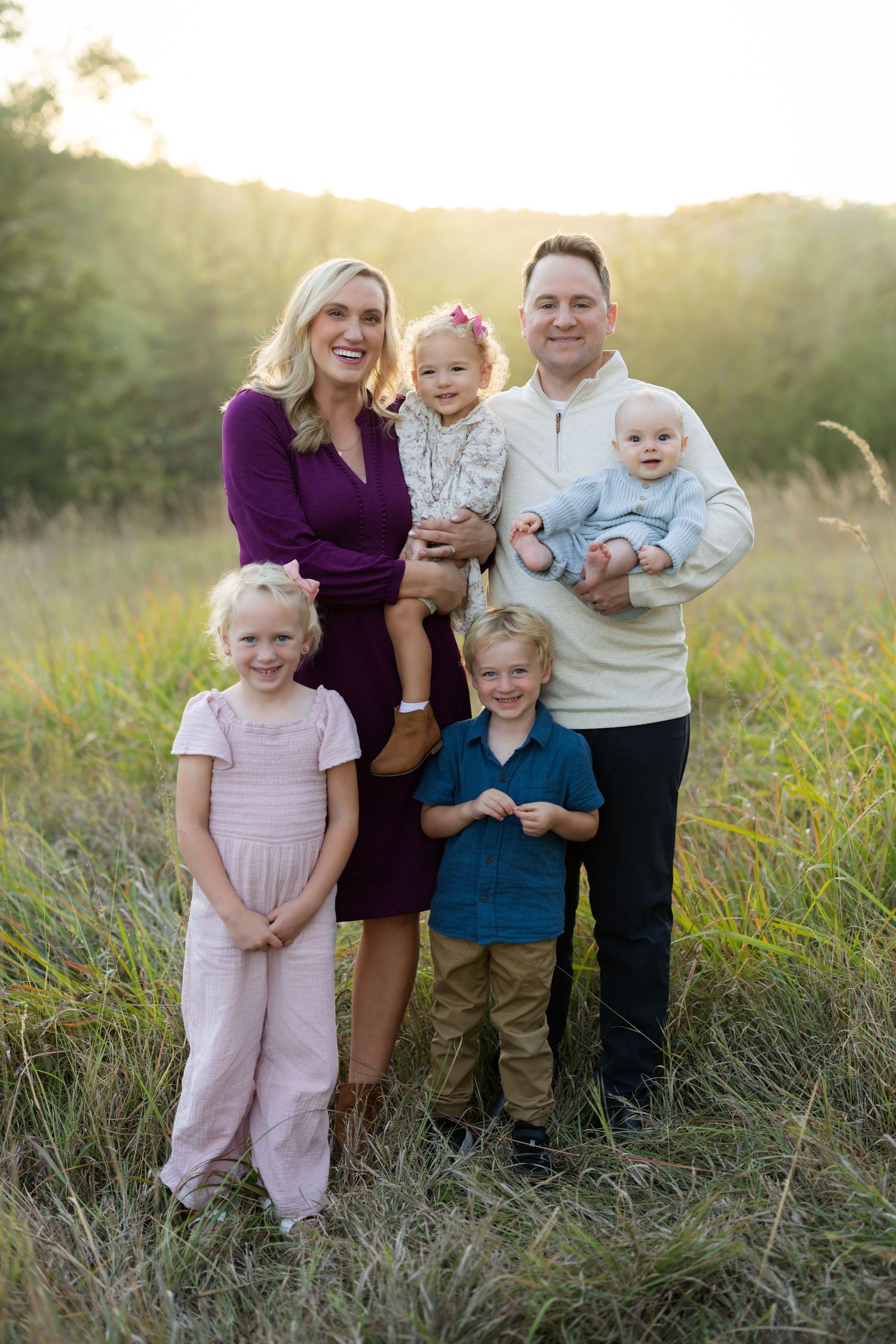 Family of six posing in a sunlit field, parents holding two toddlers while two young children stand in front.