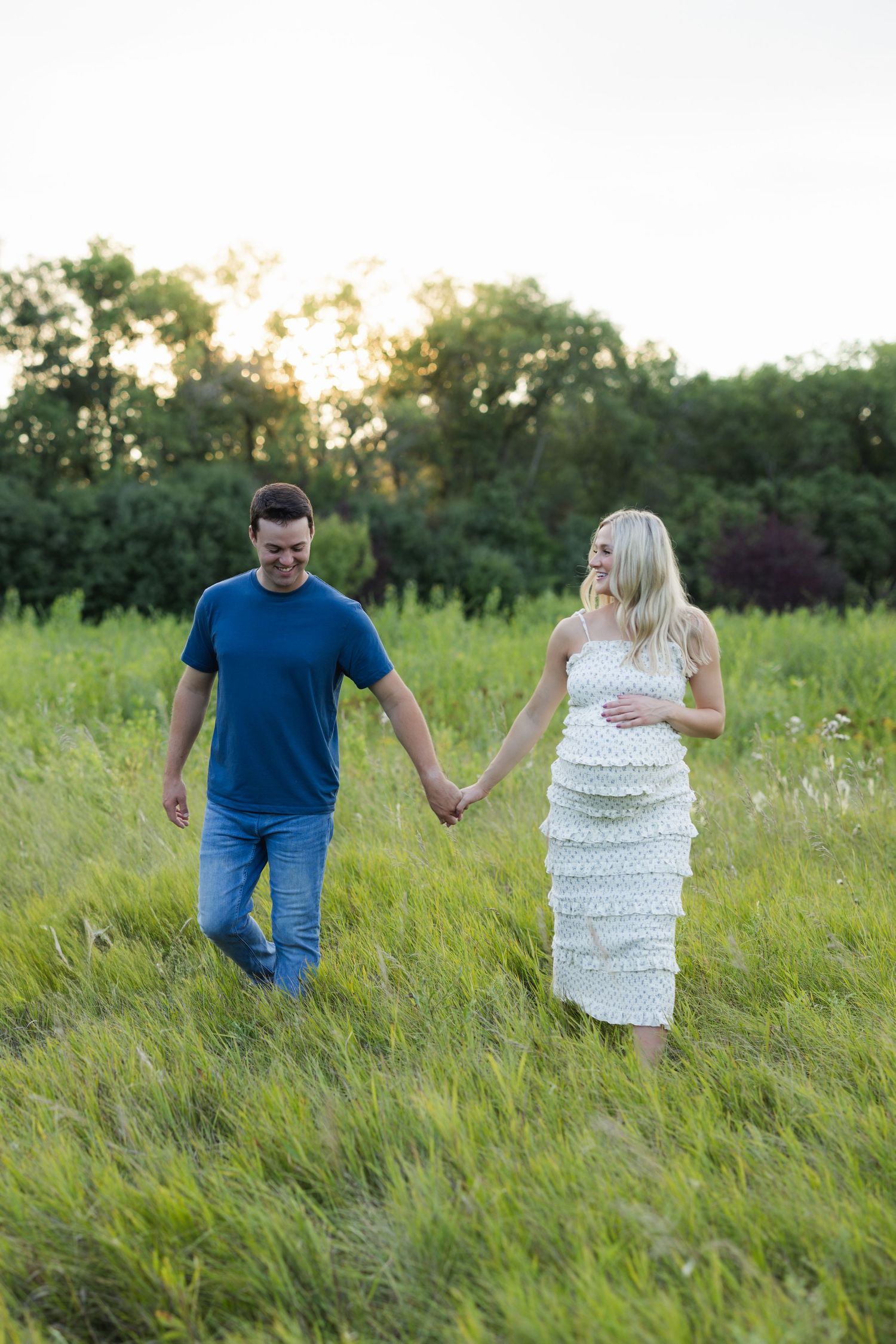 Couple holding hands walking through lush green field at sunset, woman in white dress touching baby bump.
