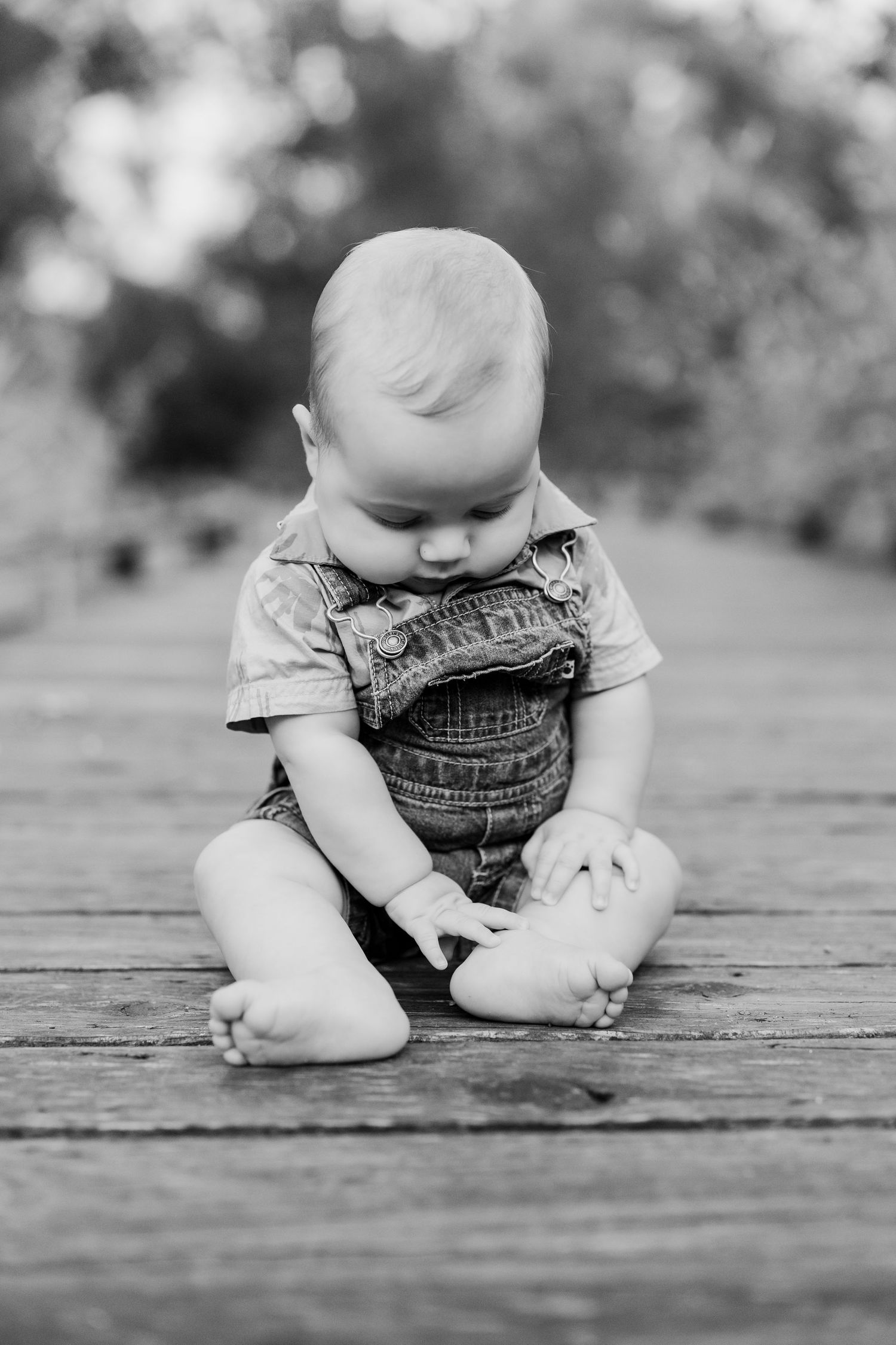Sweet black and white portrait of a baby in denim overalls sitting on wooden deck looking down.