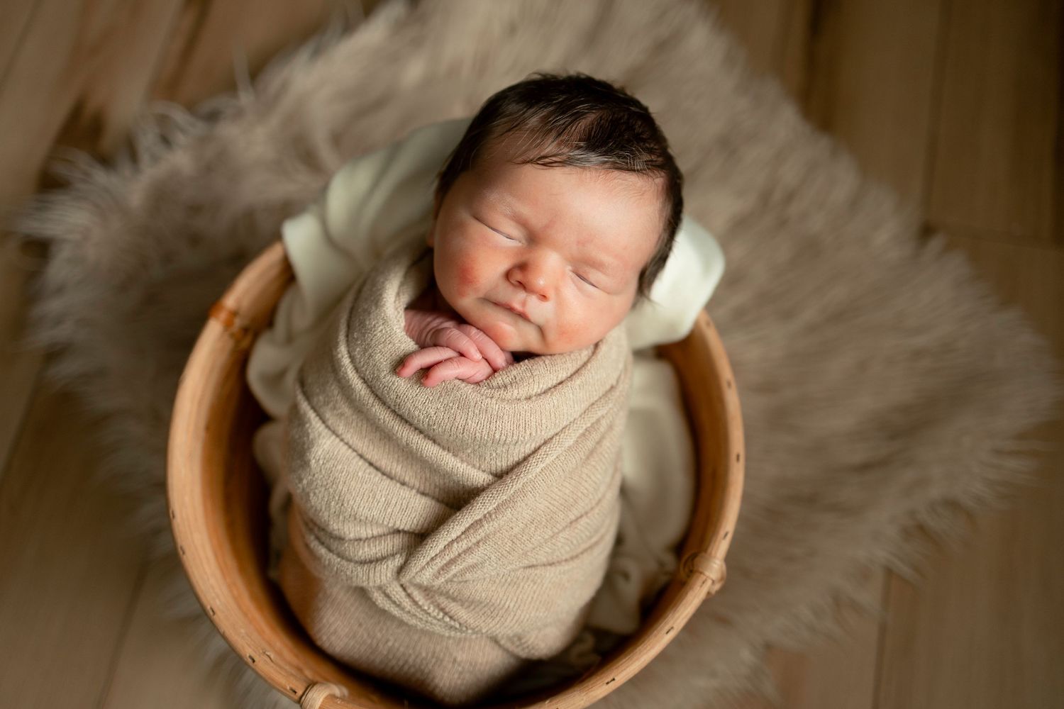 Sleeping newborn baby swaddled in beige wrap, resting in a wooden bowl on a fluffy tan fur rug.
