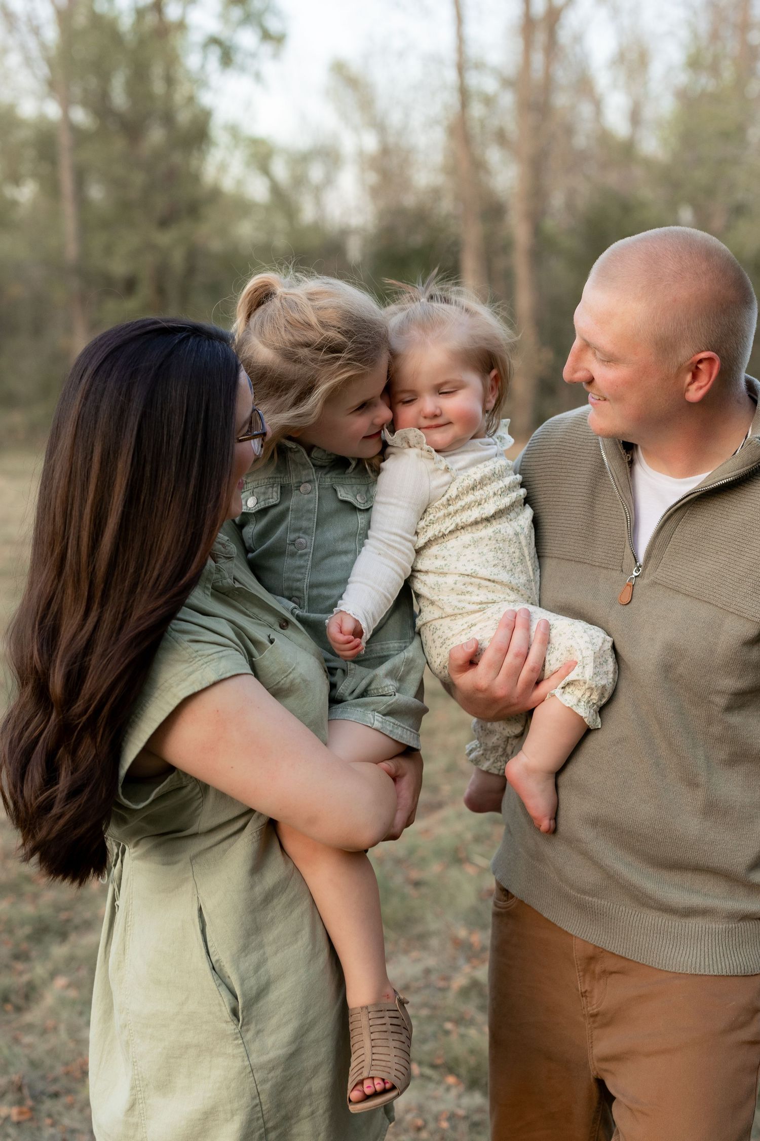 Family of four posing outdoors, parents holding two young daughters, smiling warmly in a wooded setting.