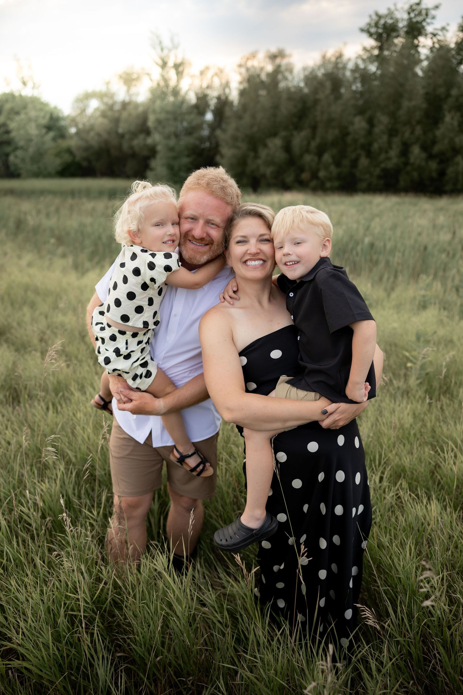 Happy family of four posing in a green meadow, parents holding two young blonde children, wearing polka dot outfits.