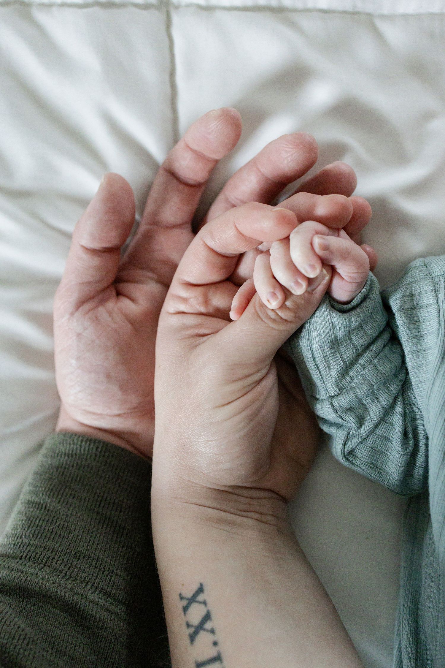 Newborn baby hand grasping parents fingers, with a Roman numeral tattoo visible on wrist, on white bedding.