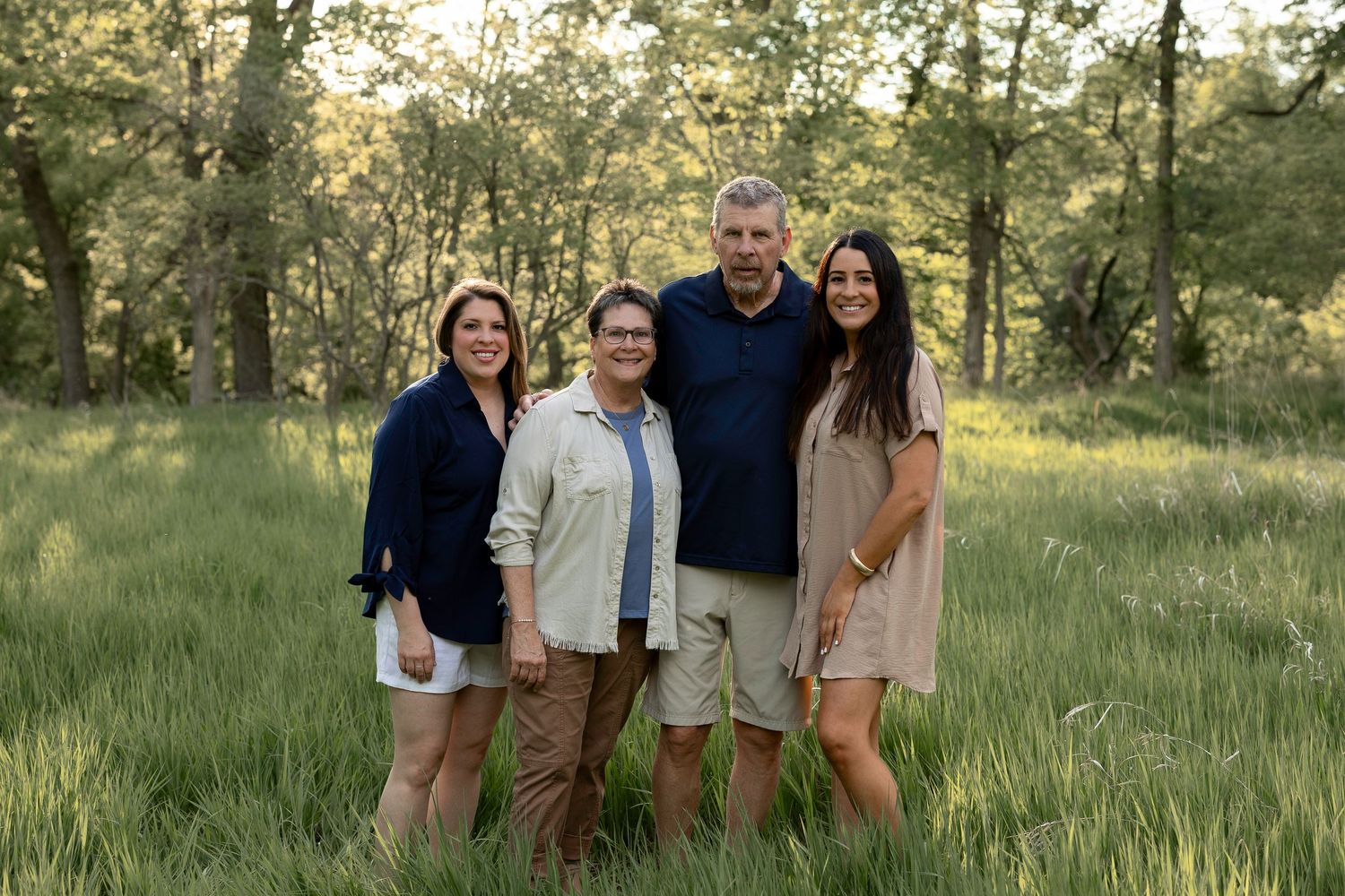 Family of four smiling together in a lush green meadow surrounded by trees during golden hour.
