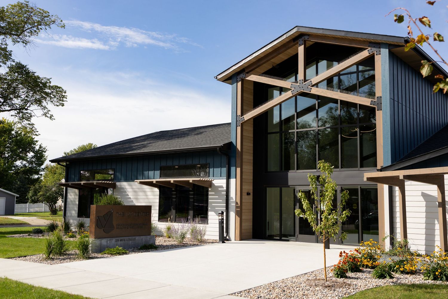Modern residential home exterior with angular architecture and large glass windows set against blue sky and landscaping.