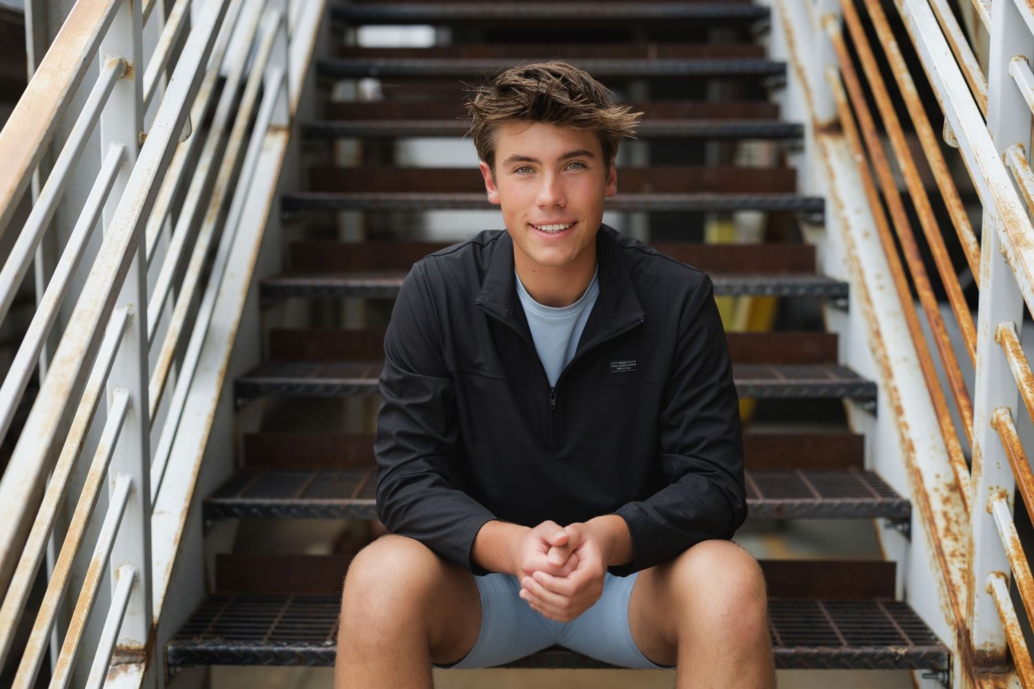 A smiling young man in a gray sweater sits casually on wooden stairs in an indoor setting.