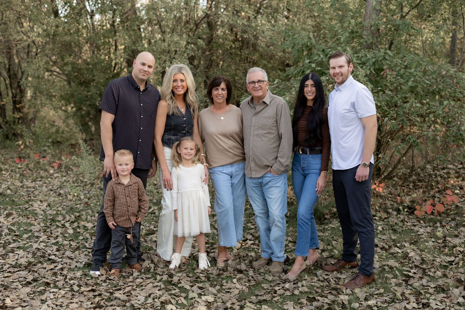 A multi-generational family group poses together outdoors in a wooded setting during a casual photo session.