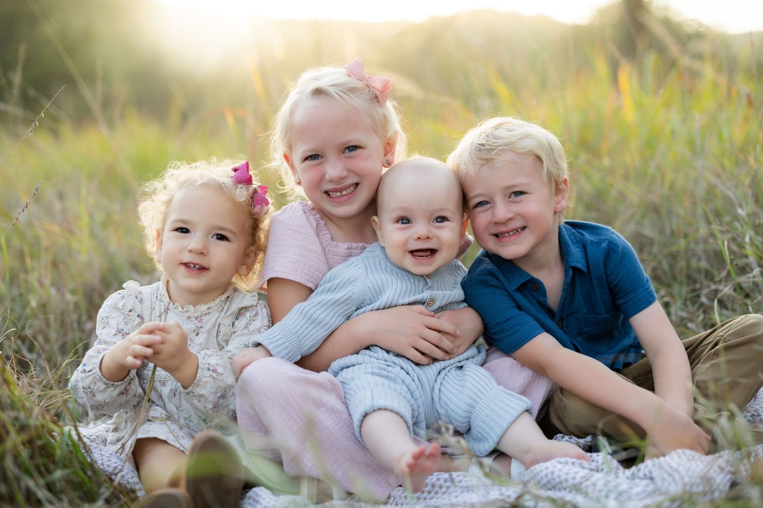 Four young blonde siblings smile joyfully while sitting together in a sunlit meadow for a family portrait.