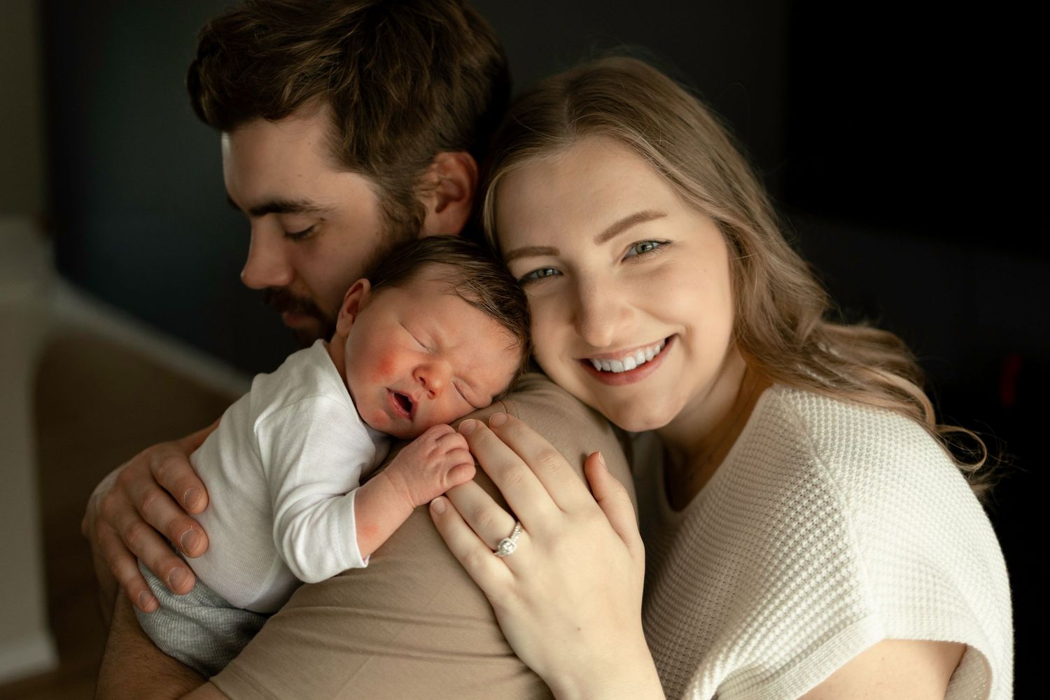 Happy couple smiling and holding their sleeping newborn baby close in a warm, intimate family portrait.