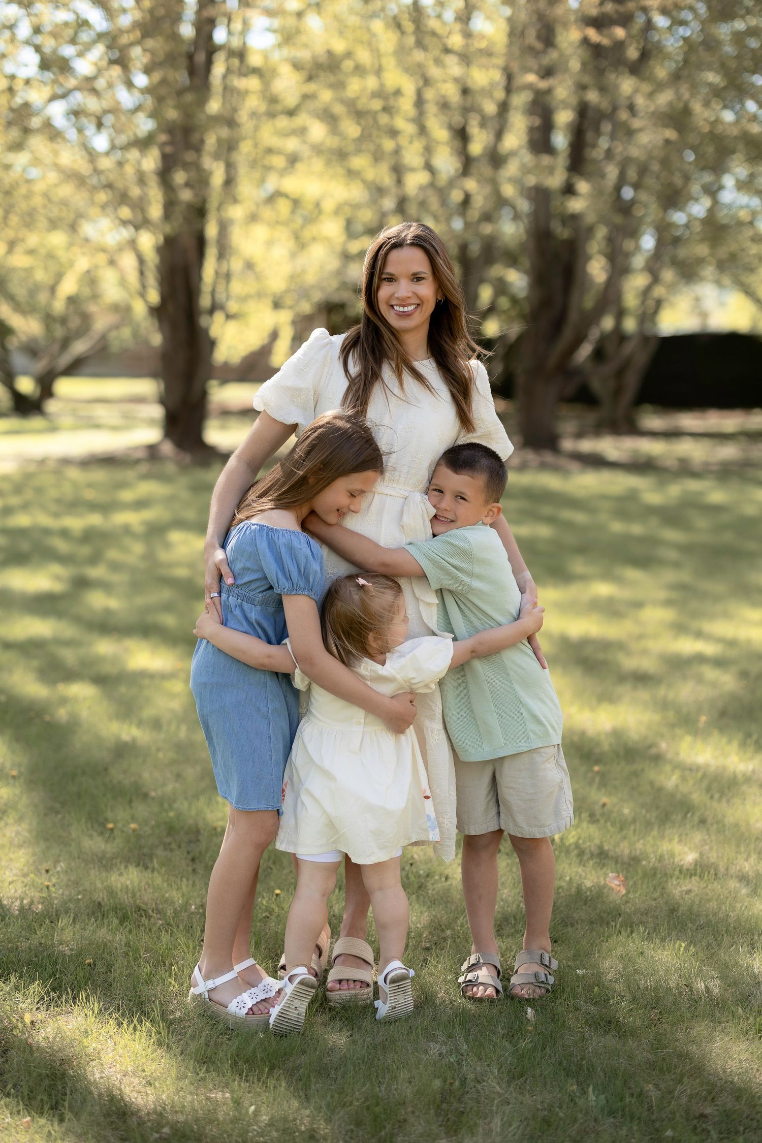 A smiling woman in a white dress hugs three young children on a green lawn surrounded by trees.