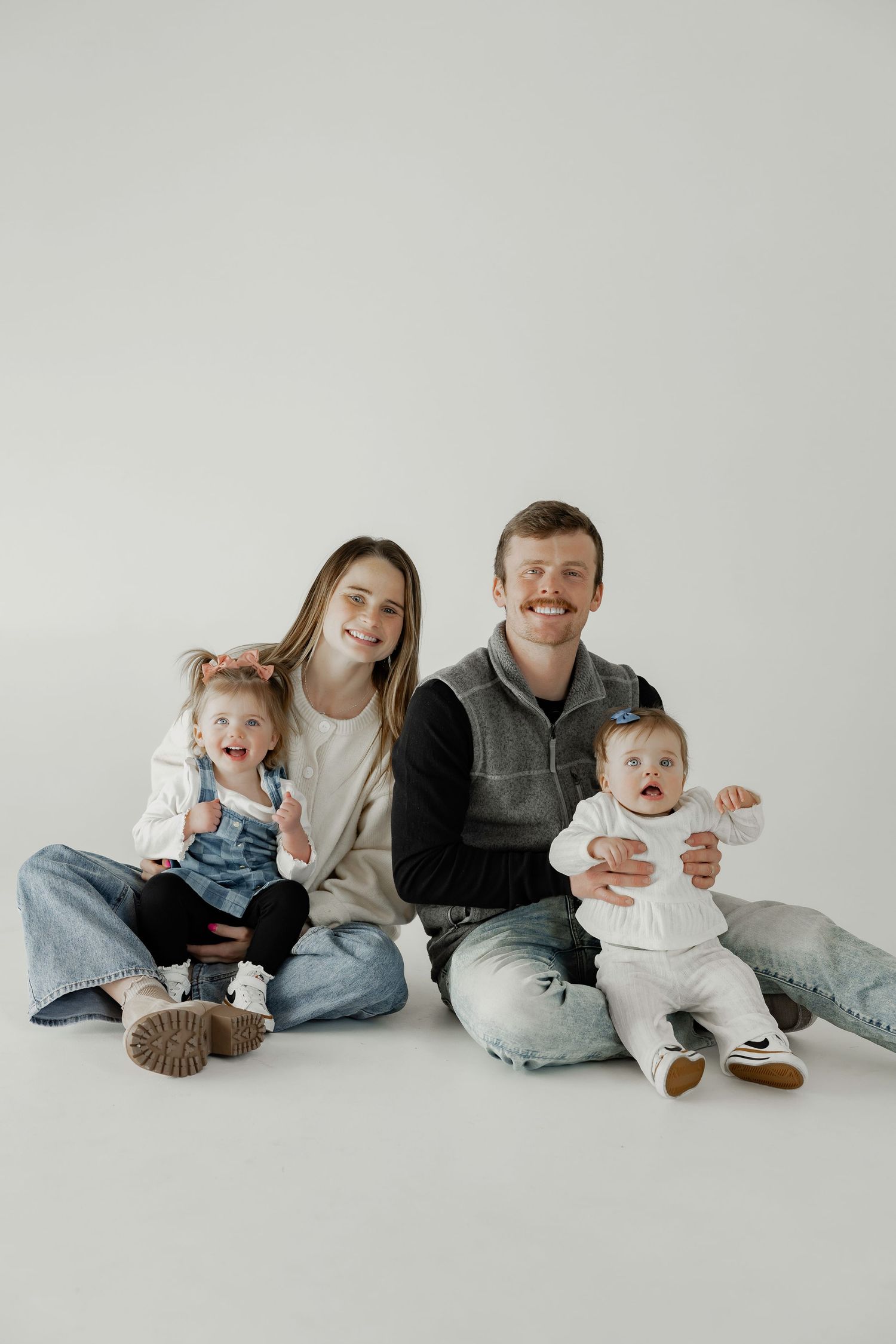 Young family with two babies sitting on white studio floor, smiling for a professional family portrait photo.