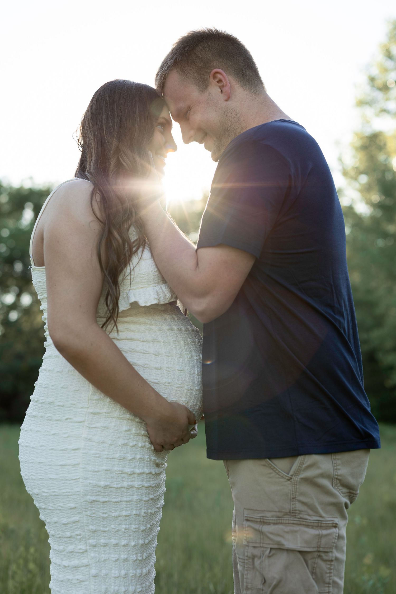 Pregnant couple touching foreheads tenderly outdoors with golden sunlight glowing between them in a green field.