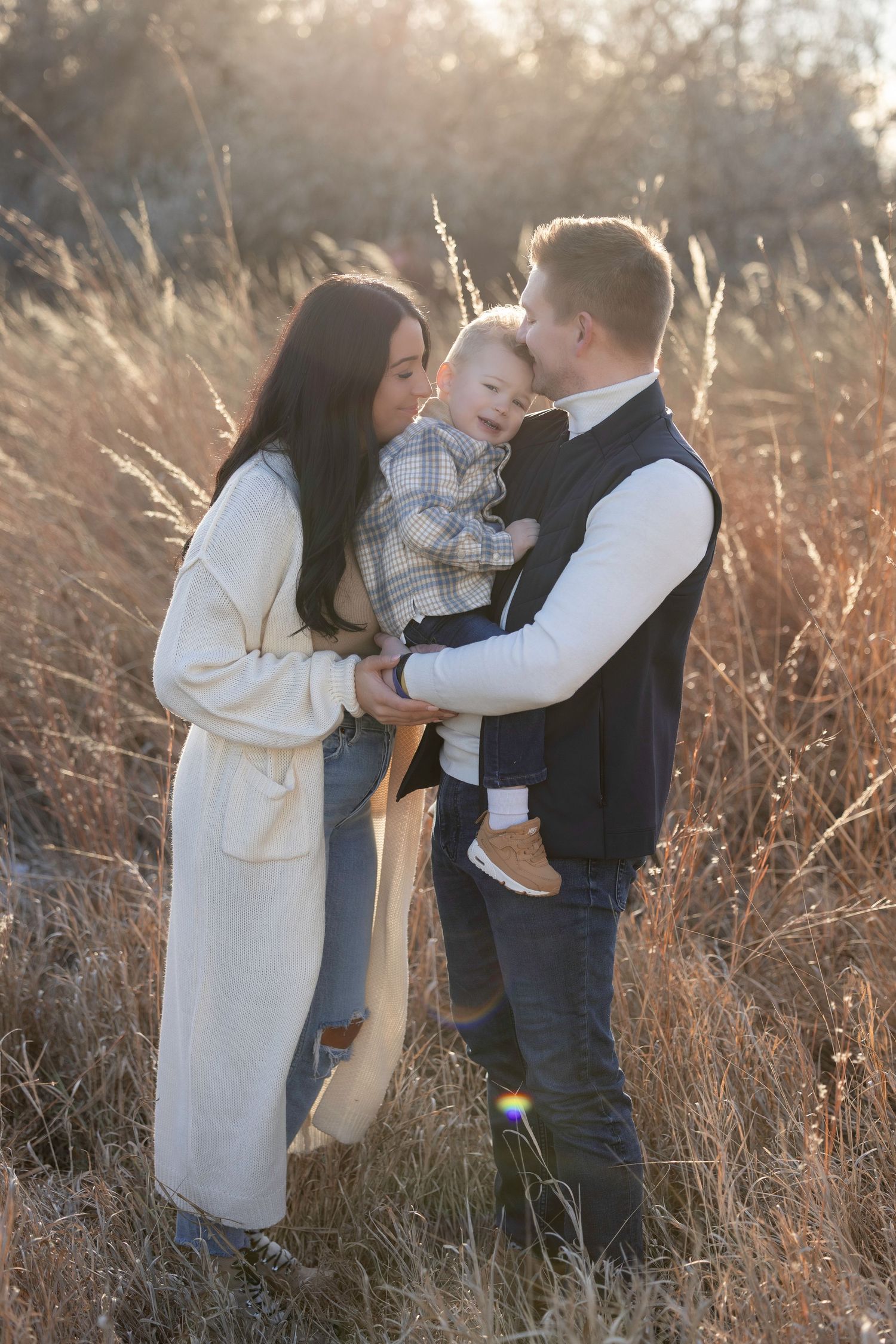 Young family with baby boy posing in golden field during sunset, parents smiling at laughing toddler.
