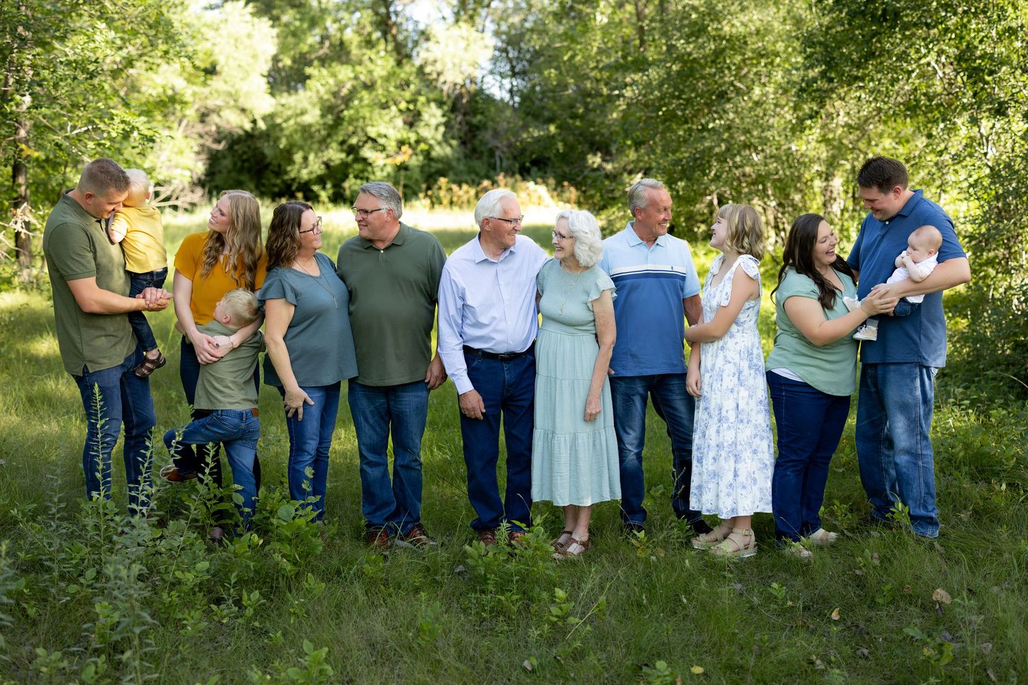 A group of people in casual attire stand in a line against a backdrop of green trees and foliage.