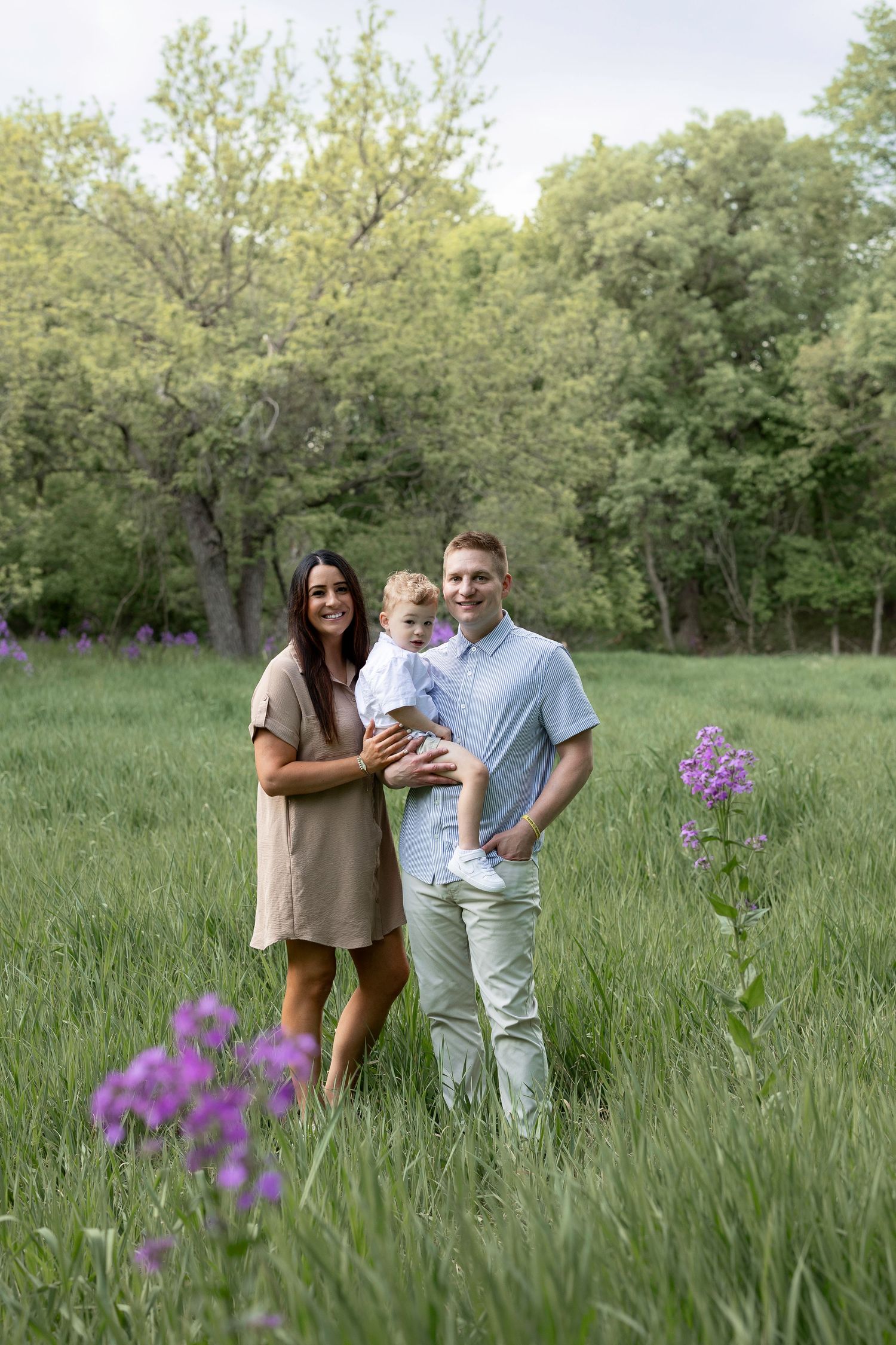 Family of three posing in a lush green meadow with purple wildflowers and trees in the background.