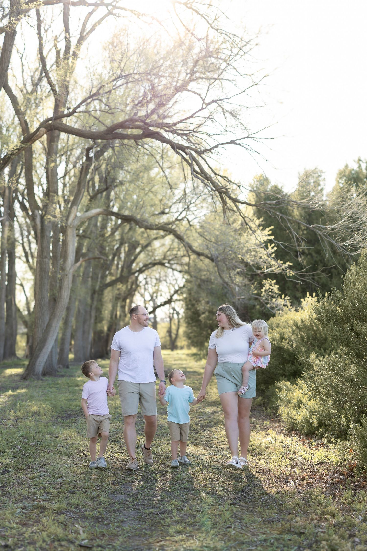 Family of five walking on a sunny wooded path, parents and three young children dressed in light casual summer outfits.