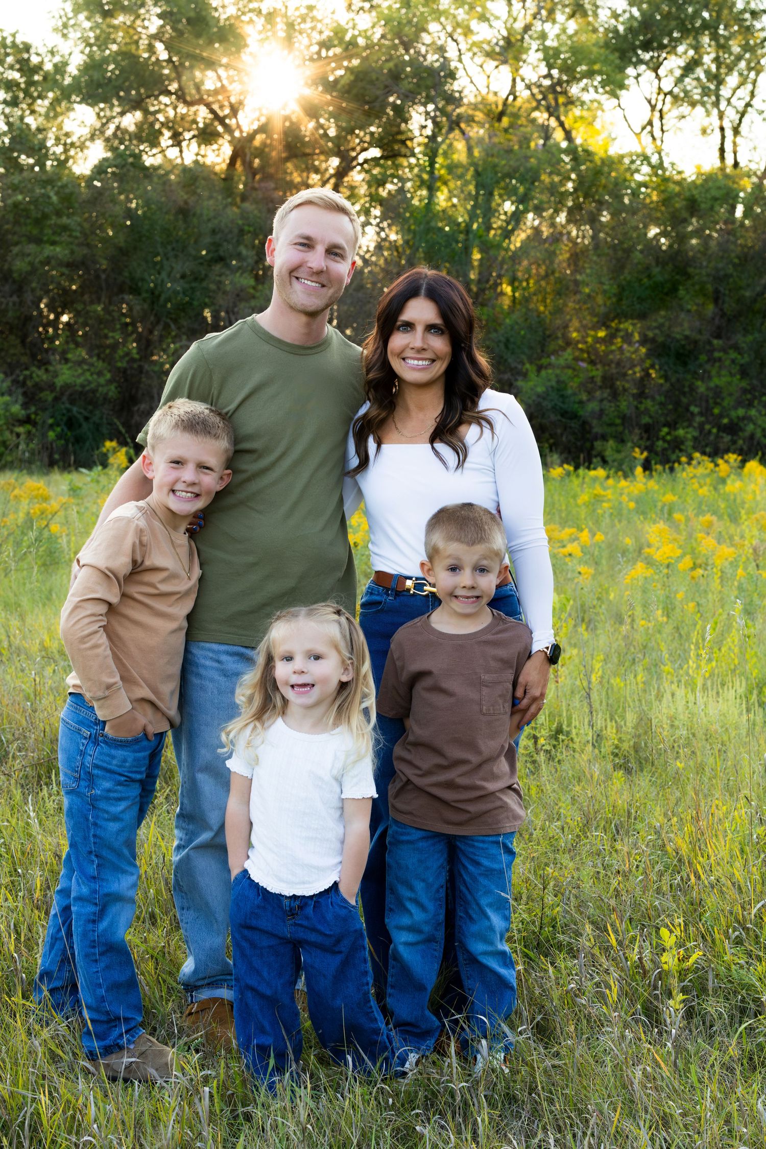 A family poses together in a field of yellow wildflowers during golden hour with sunlight streaming through trees.