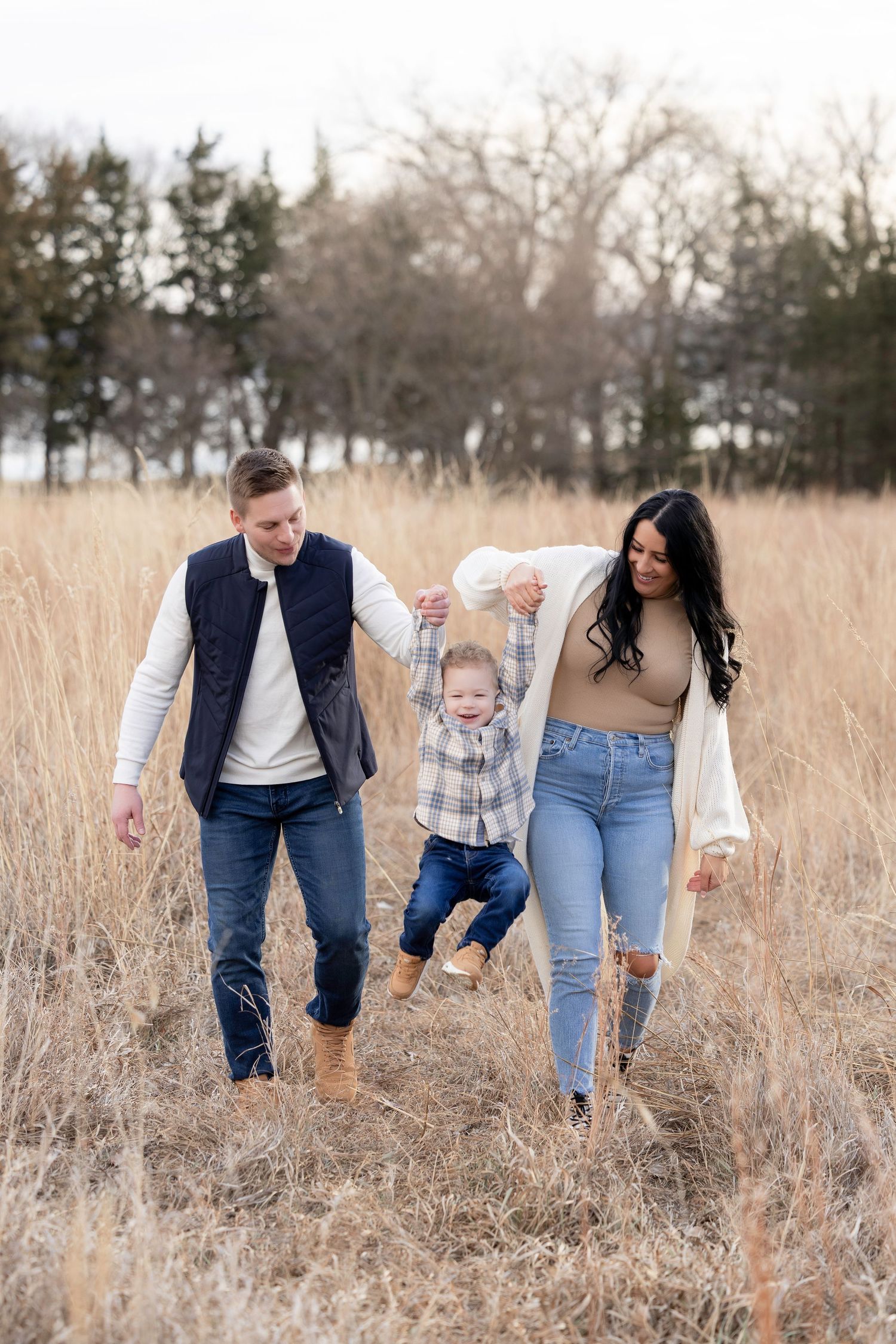 A happy family of three swings their toddler through a golden field, wearing casual fall outfits outdoors.