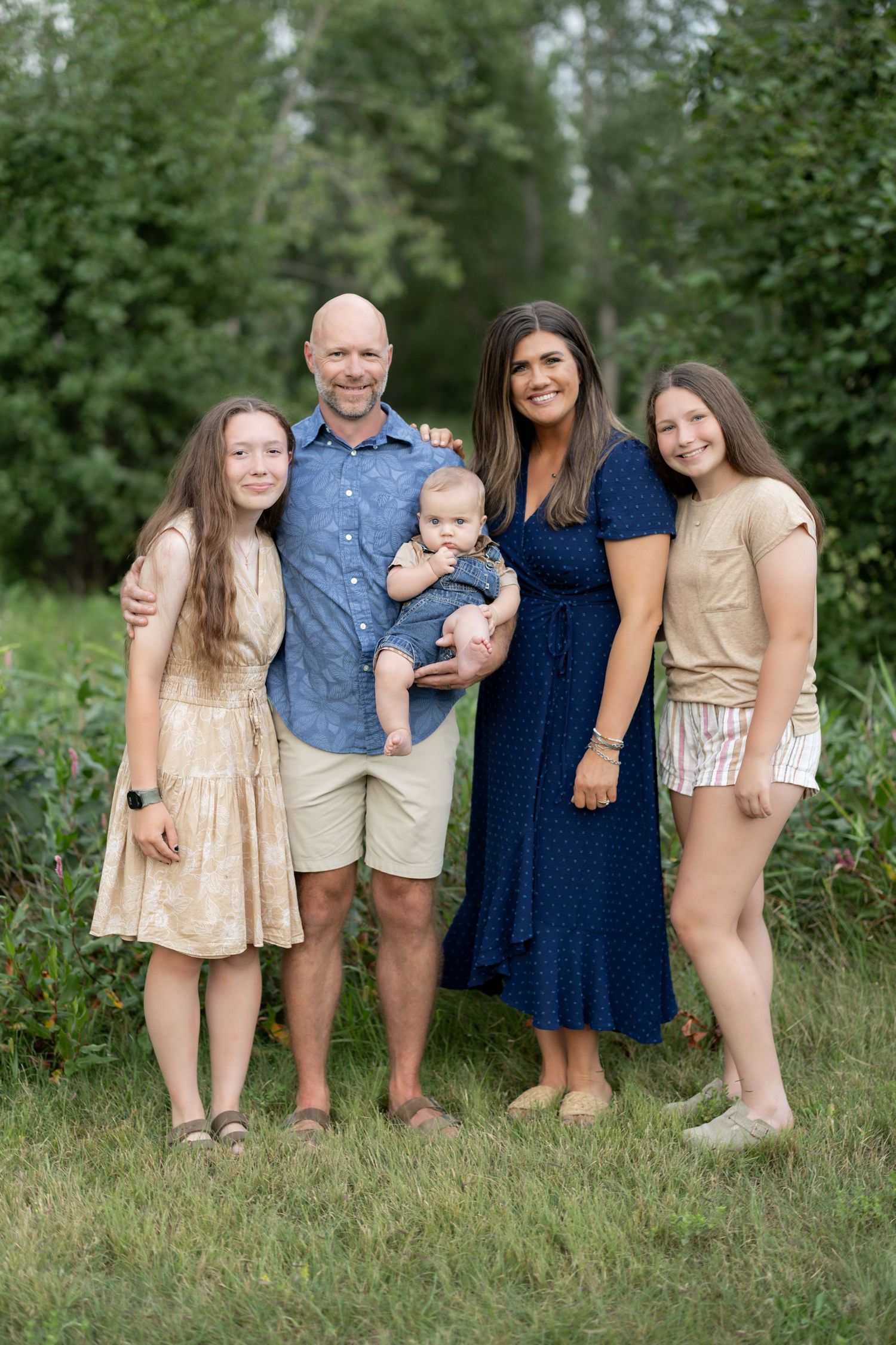 A family stands together outdoors on grass with trees in the background wearing casual summer clothing.