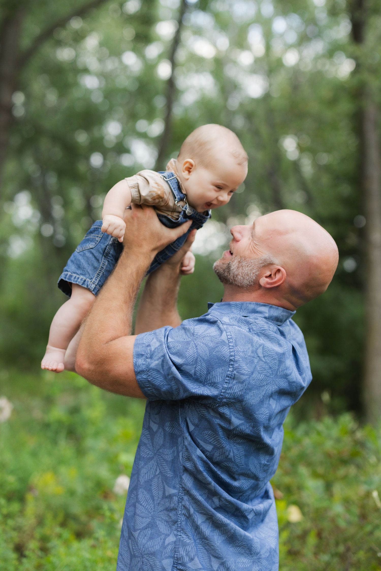 Bald father lifting smiling baby in denim outfit outdoors in lush green park, sharing a joyful moment together.