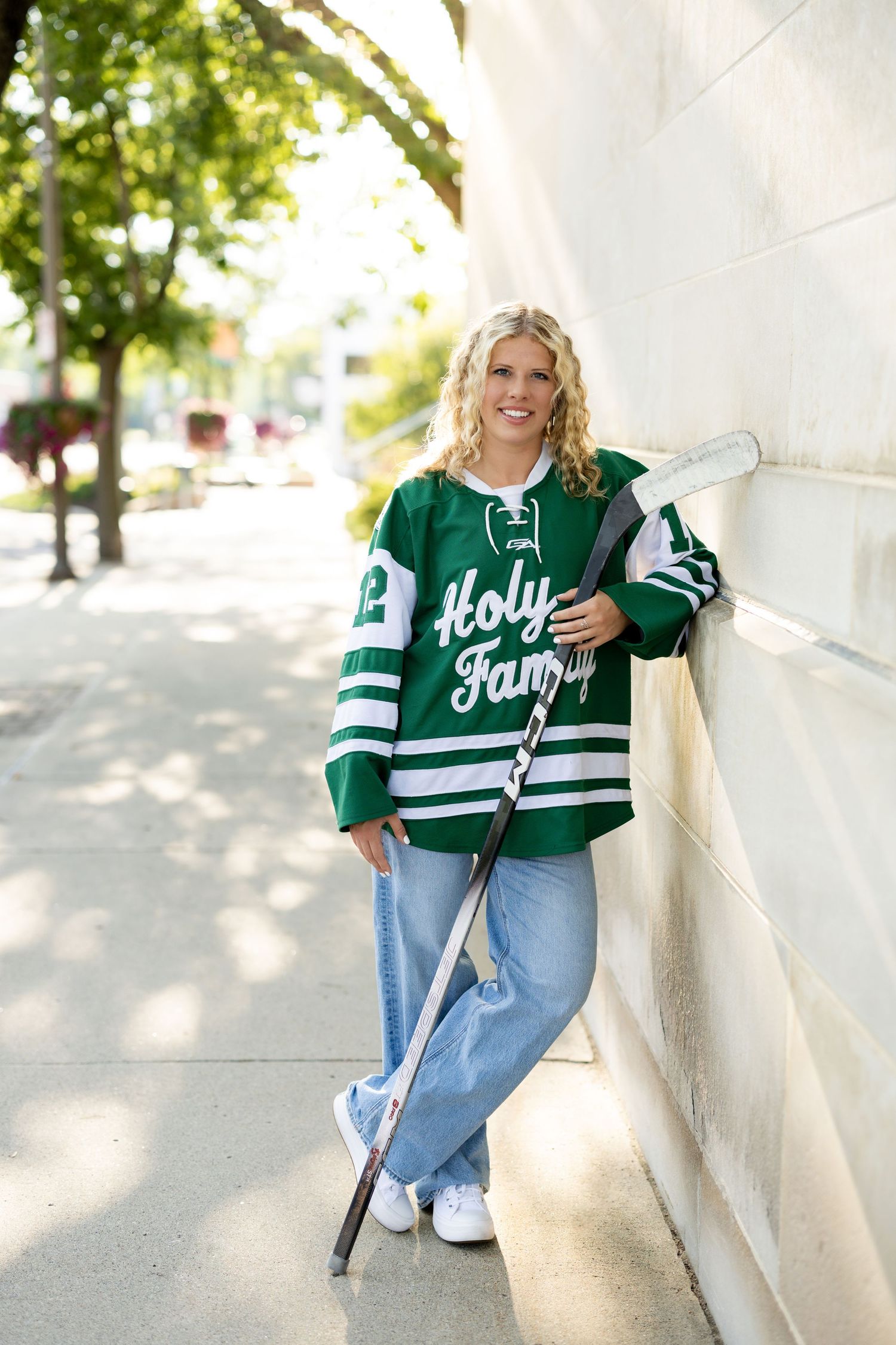 Person wearing green hockey jersey and jeans poses against white wall in outdoor setting with trees in background.