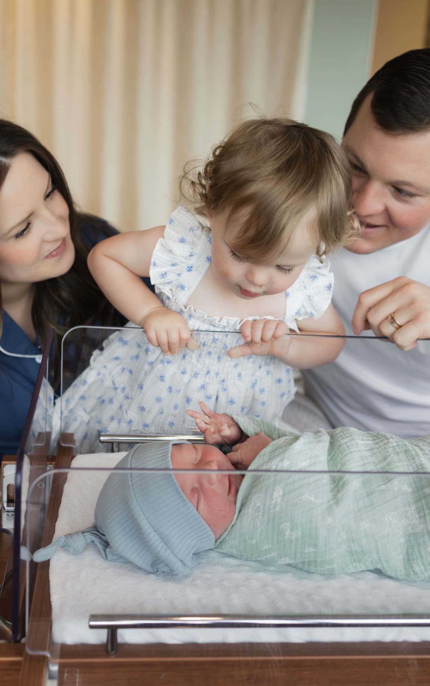Family with toddler girl admiring newborn baby in hospital bassinet, parents smiling with joy.