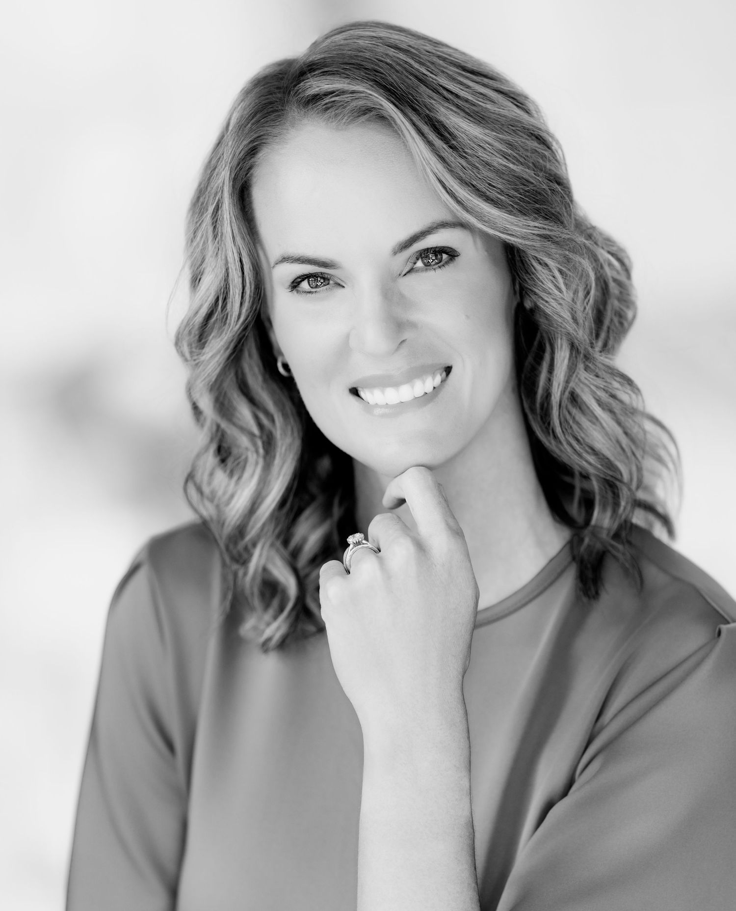 Black and white portrait of a smiling woman with wavy shoulder-length hair resting her chin on her hand, wearing a ring and a blouse.