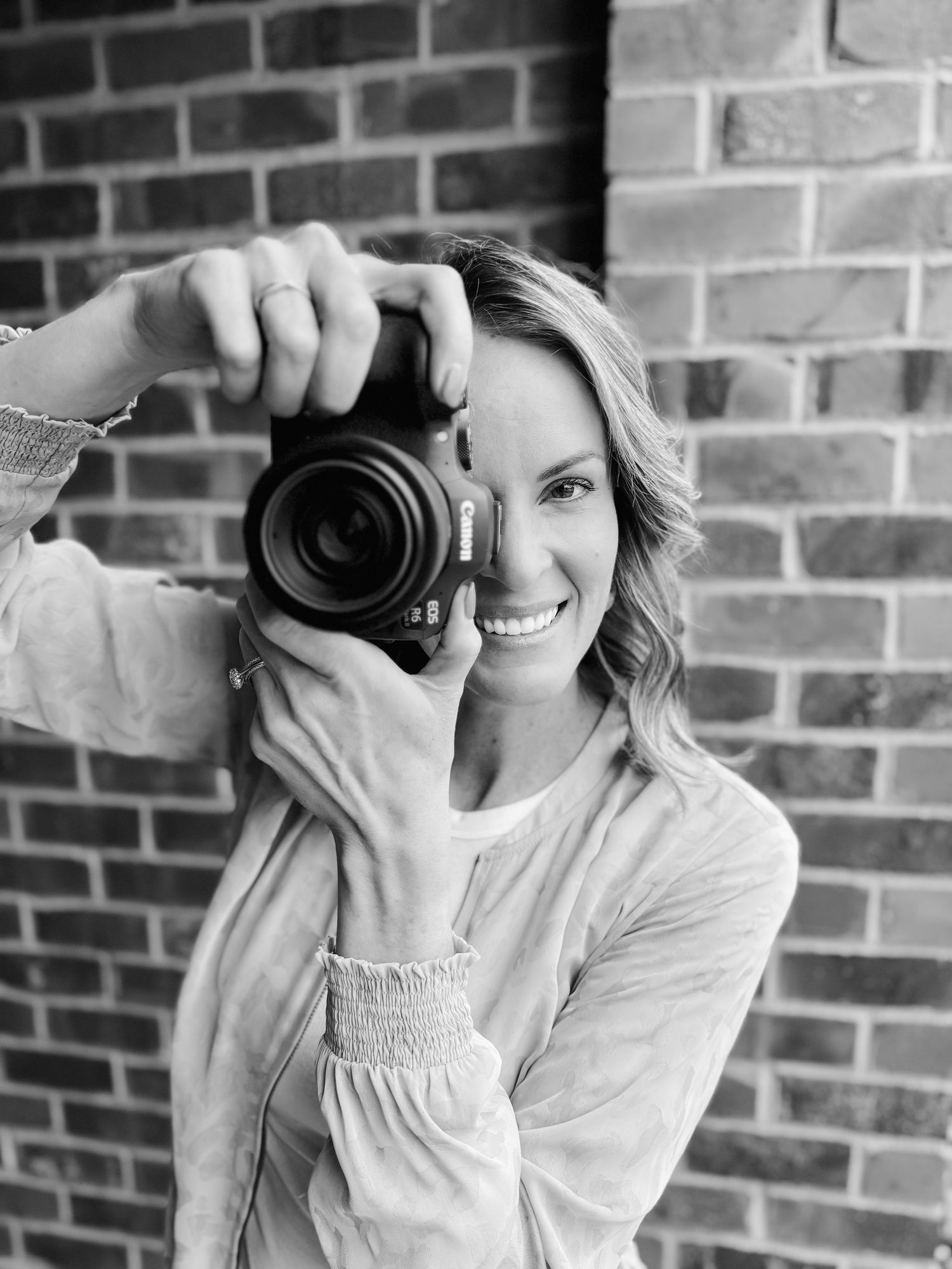 Black and white portrait of someone holding a professional camera while wearing a light colored sweater against a brick wall.