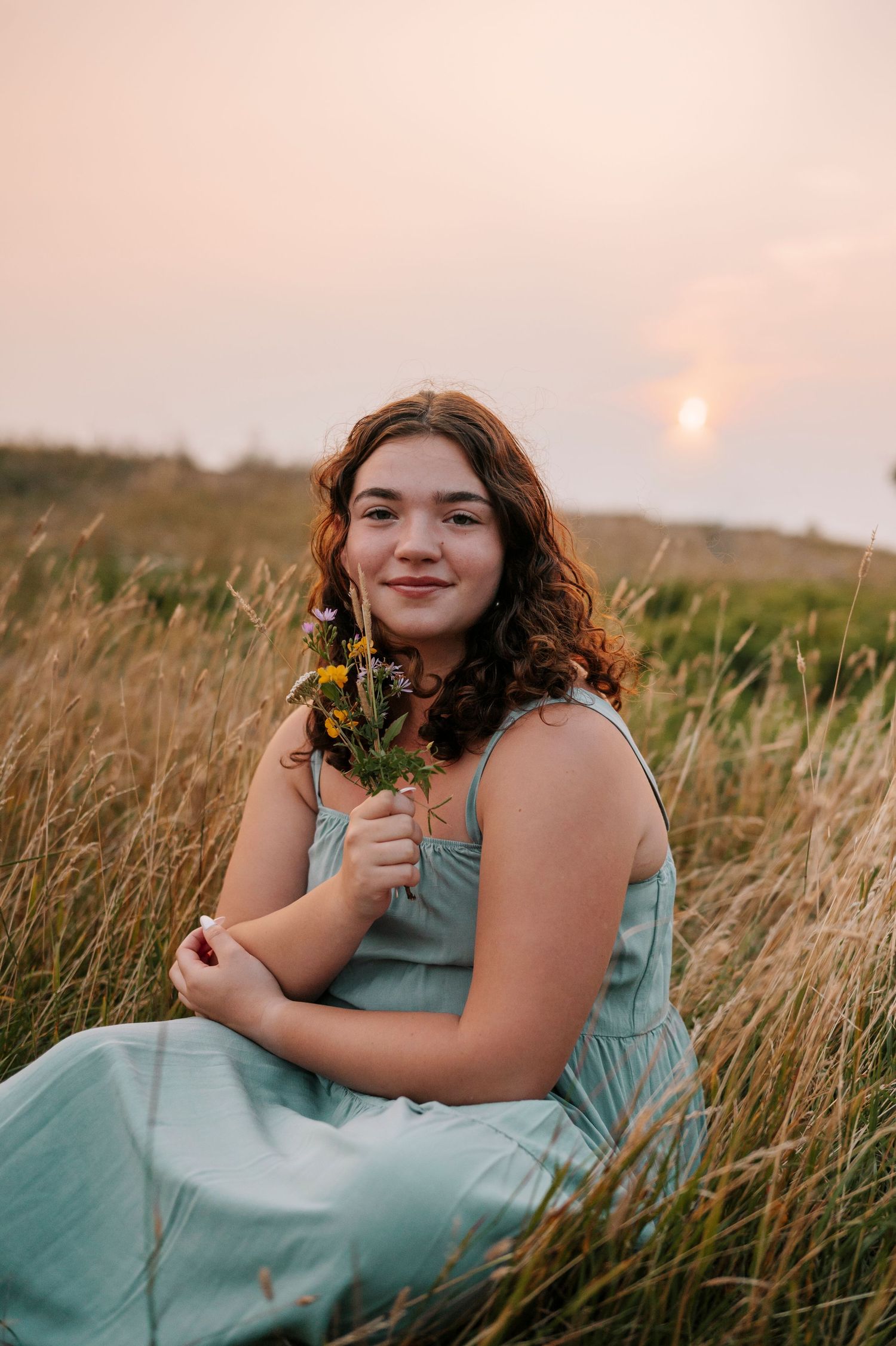 A high school senior on top of Brockway mountain holding wildflowers with the rising sun behind her