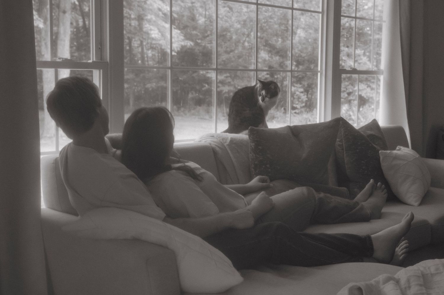 A black and white photo of a couple laying on the couch with their cat sitting in front of them